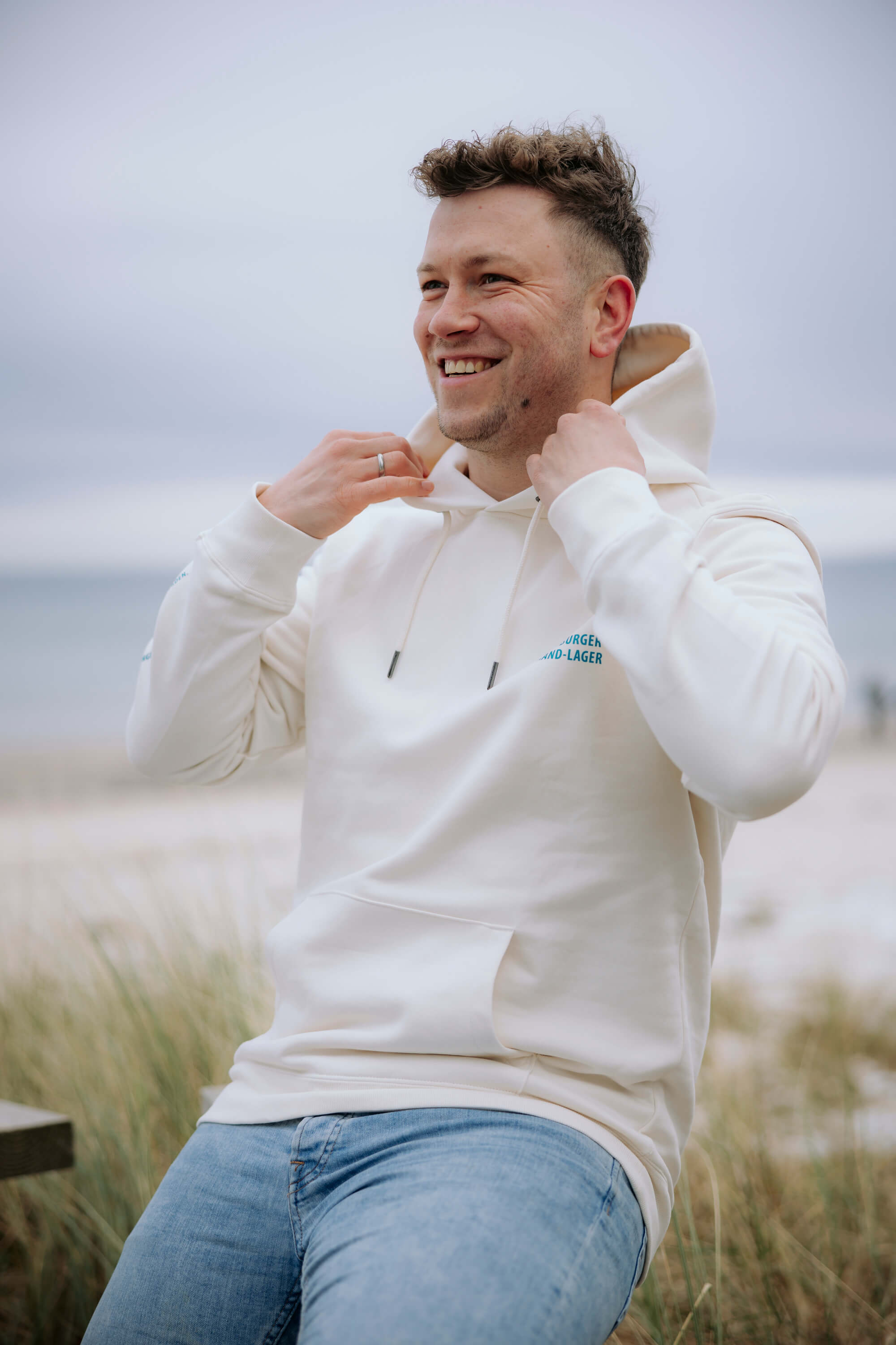 Mann trägt einen beigefarbenen Hoodie mit der türkisfarbenen Aufschrift „FLENSBURGER STRAND-LAGER“. Er hält die Kapuze mit beiden Händen und lächelt. Im Hintergrund ist ein strandähnliches Umfeld.