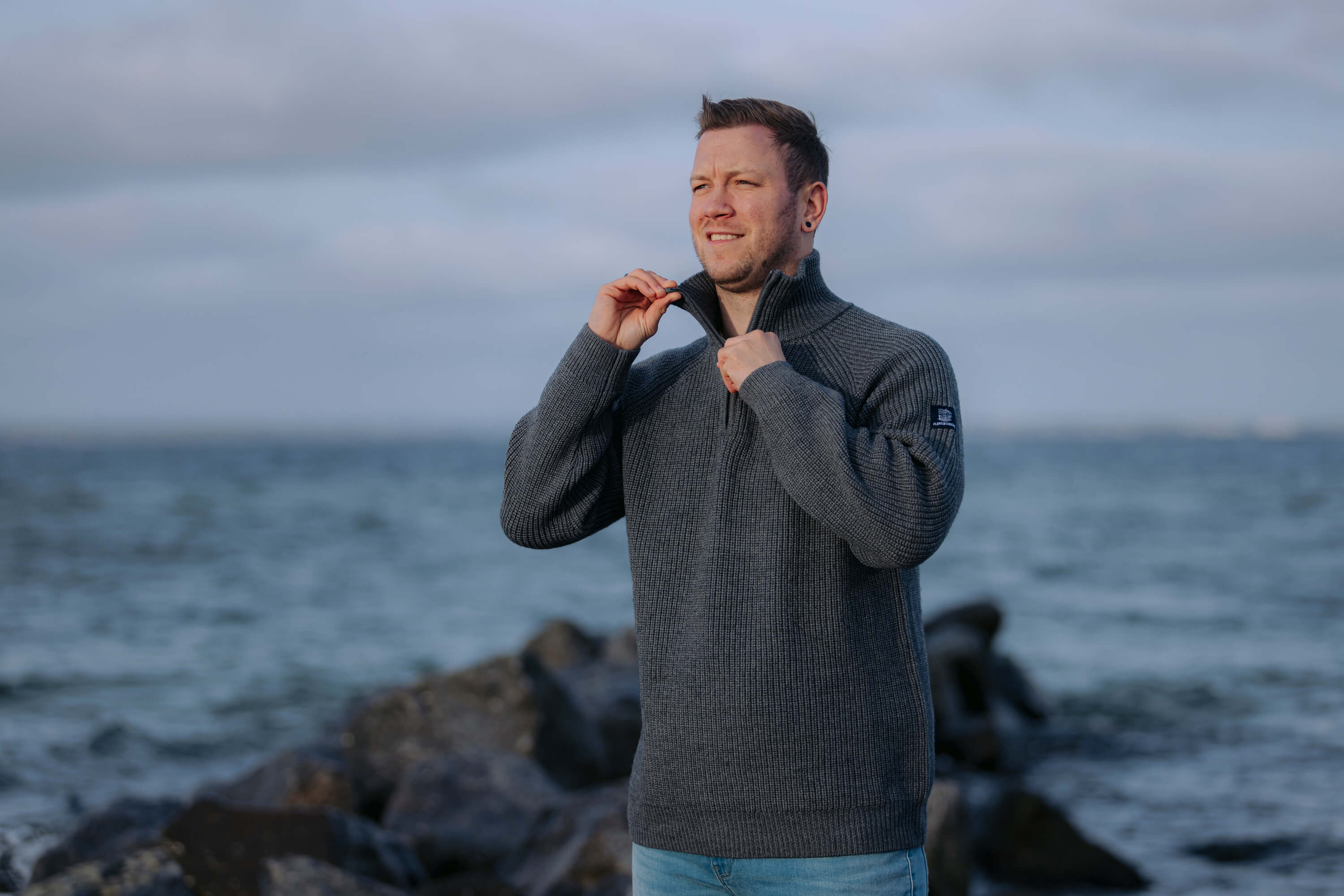 Mann steht am Meer auf Steinen und schließt den Half-Zip-Reißverschluss seines anthrazitfarbenen Strickpullovers "Tilda". Er trägt hellblaue Jeans und blickt in die Ferne bei bewölktem Himmel.