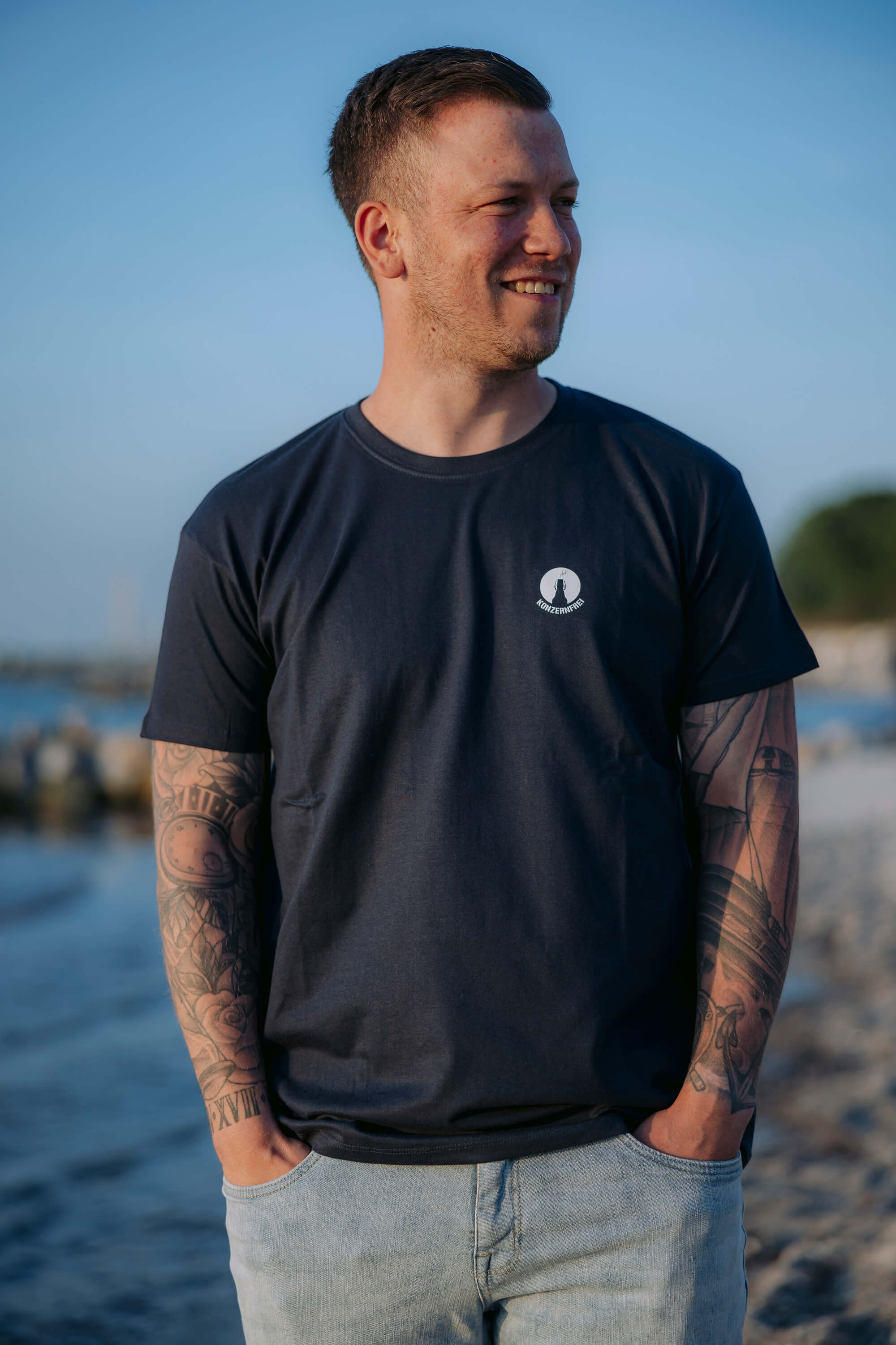 Ein Mann mit Tattoos an beiden Armen trägt ein blaues Kurzarm-T-Shirt mit kleinem weißen Logo „KONZERNFREI“ auf der linken Brustseite. Er steht am Strand und lächelt.