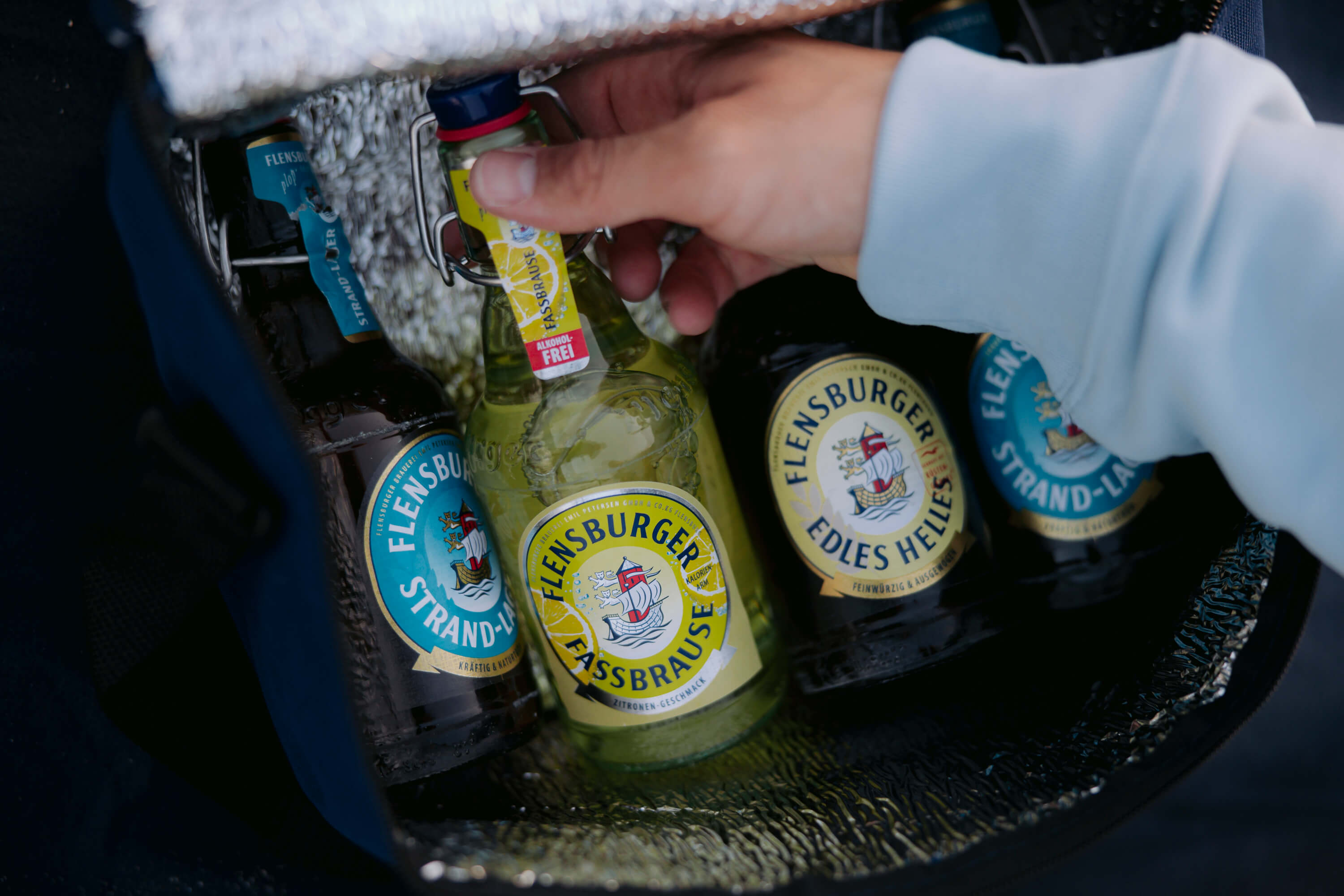 Ein Hand greift nach einer Flasche Flensburger Fassbrause in einer blauen Kühltasche. Die Flasche ist zwischen weiteren Flensburger Getränken platziert.