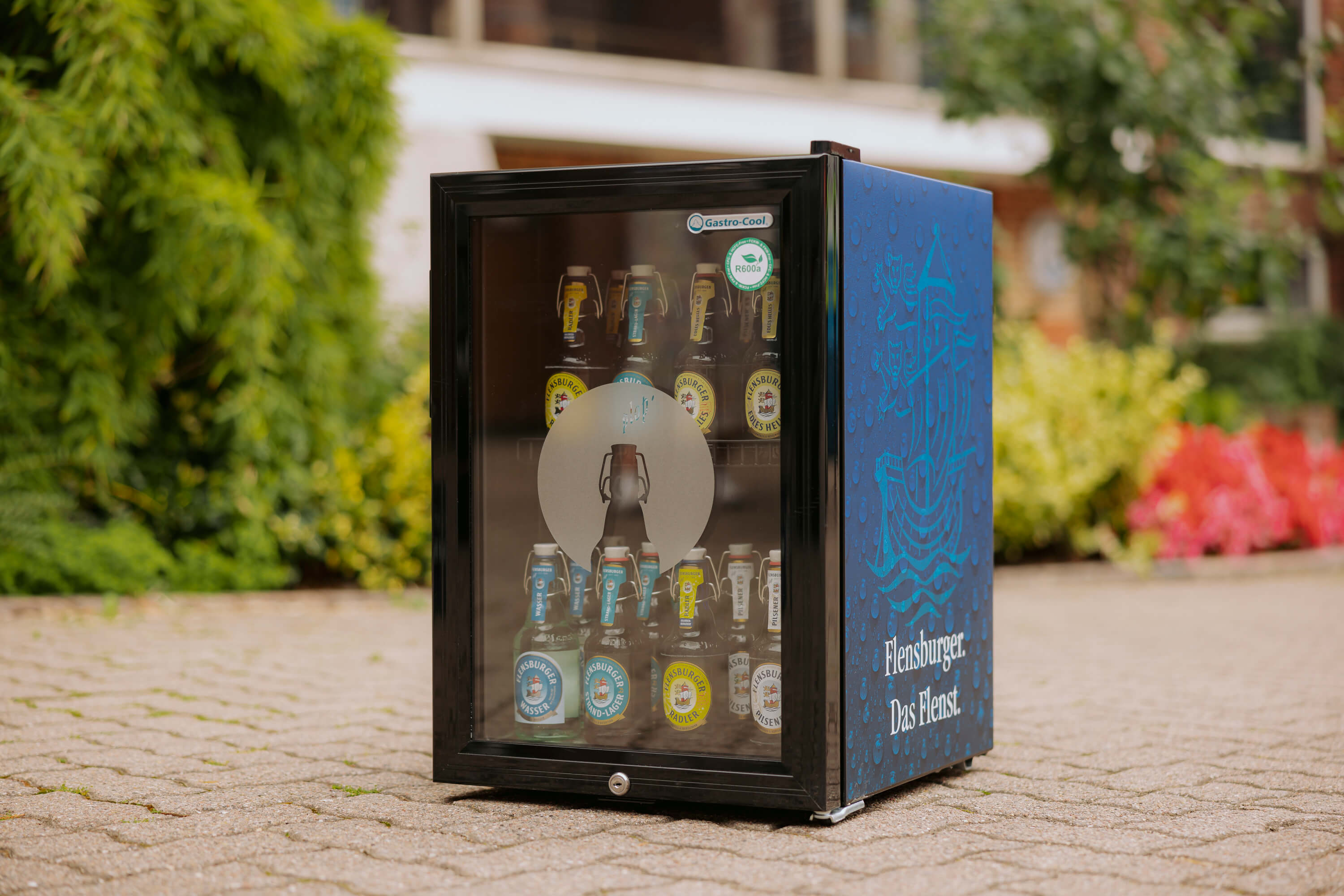 Ein navyblauer Tresenkühlschrank mit Glasfront und Schloss zeigt verschiedene Flensburger Bierflaschen. Auf der Seite steht „Flensburger. Das Flenst.“. Der Kühlschrank steht draußen auf gepflastertem Boden.