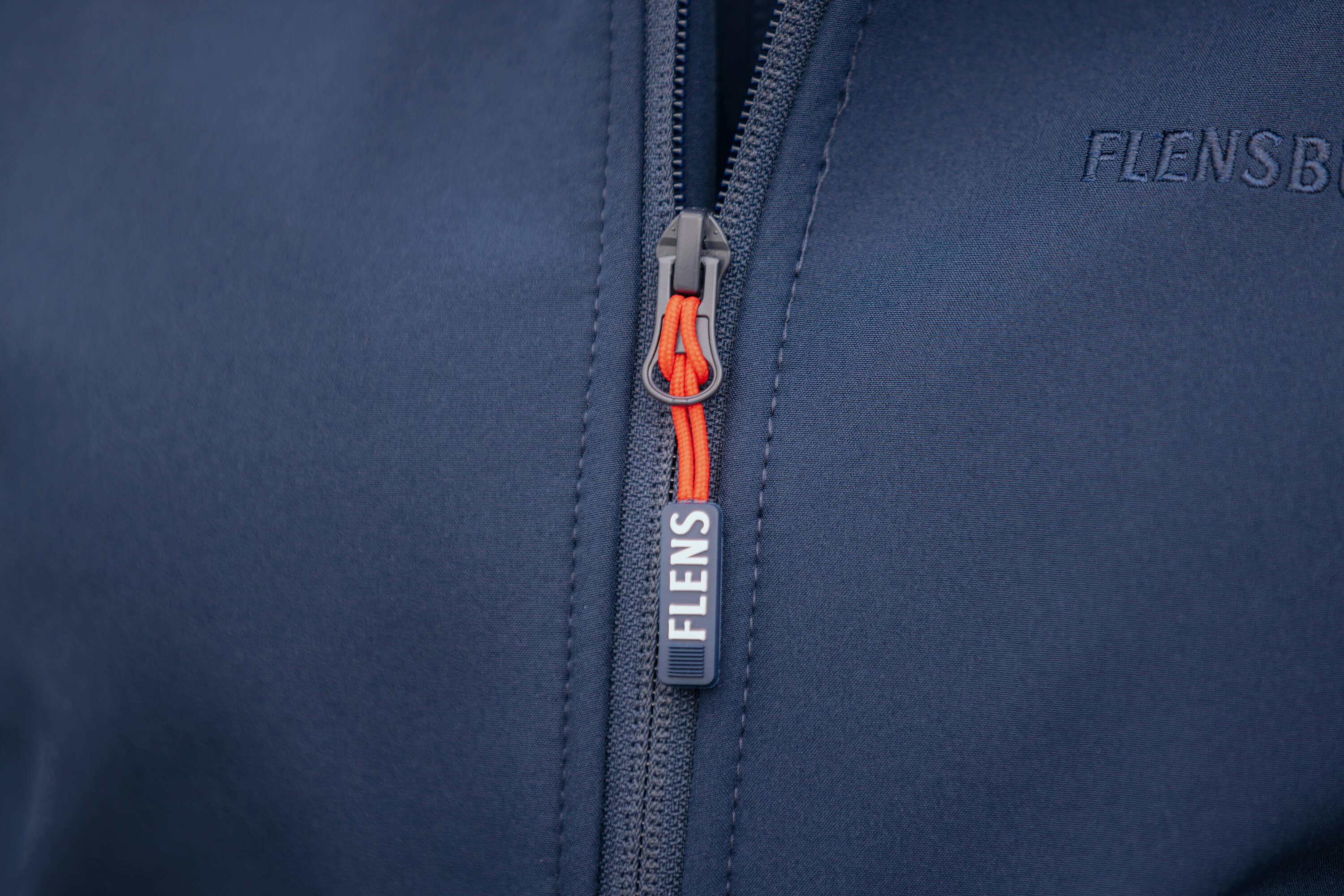 Detailaufnahme eines navyblauen Softshelljacken-Reißverschlusses mit orangem Zipper-Anhänger und dem weißen Schriftzug „FLENS“. Auf der Brust ist der Schriftzug "FLENSBURGER" in Navy gestickt.