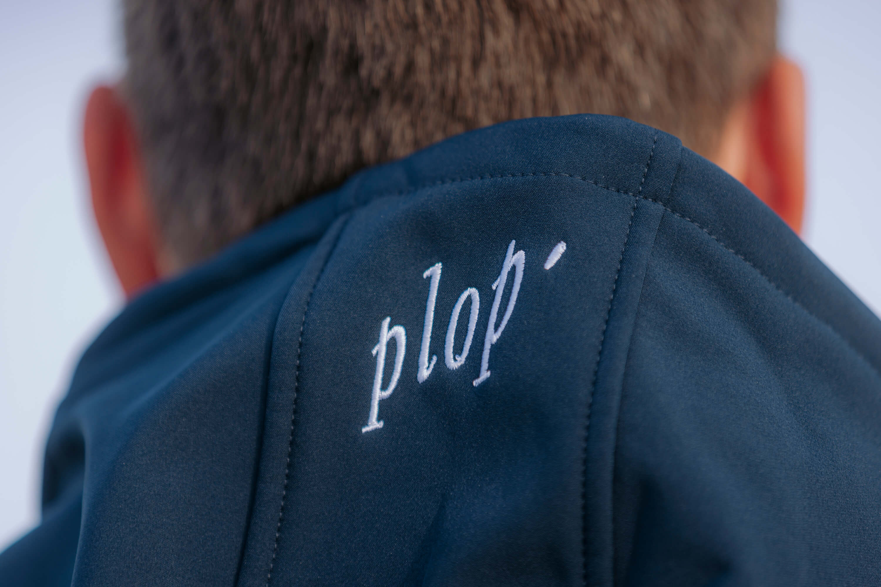 Nahaufnahme des Kragens einer dunkelblauen Softshelljacke mit weiß gesticktem Schriftzug "plop'". Im Hintergrund ist der Hinterkopf einer Person mit kurzen Haaren zu sehen.