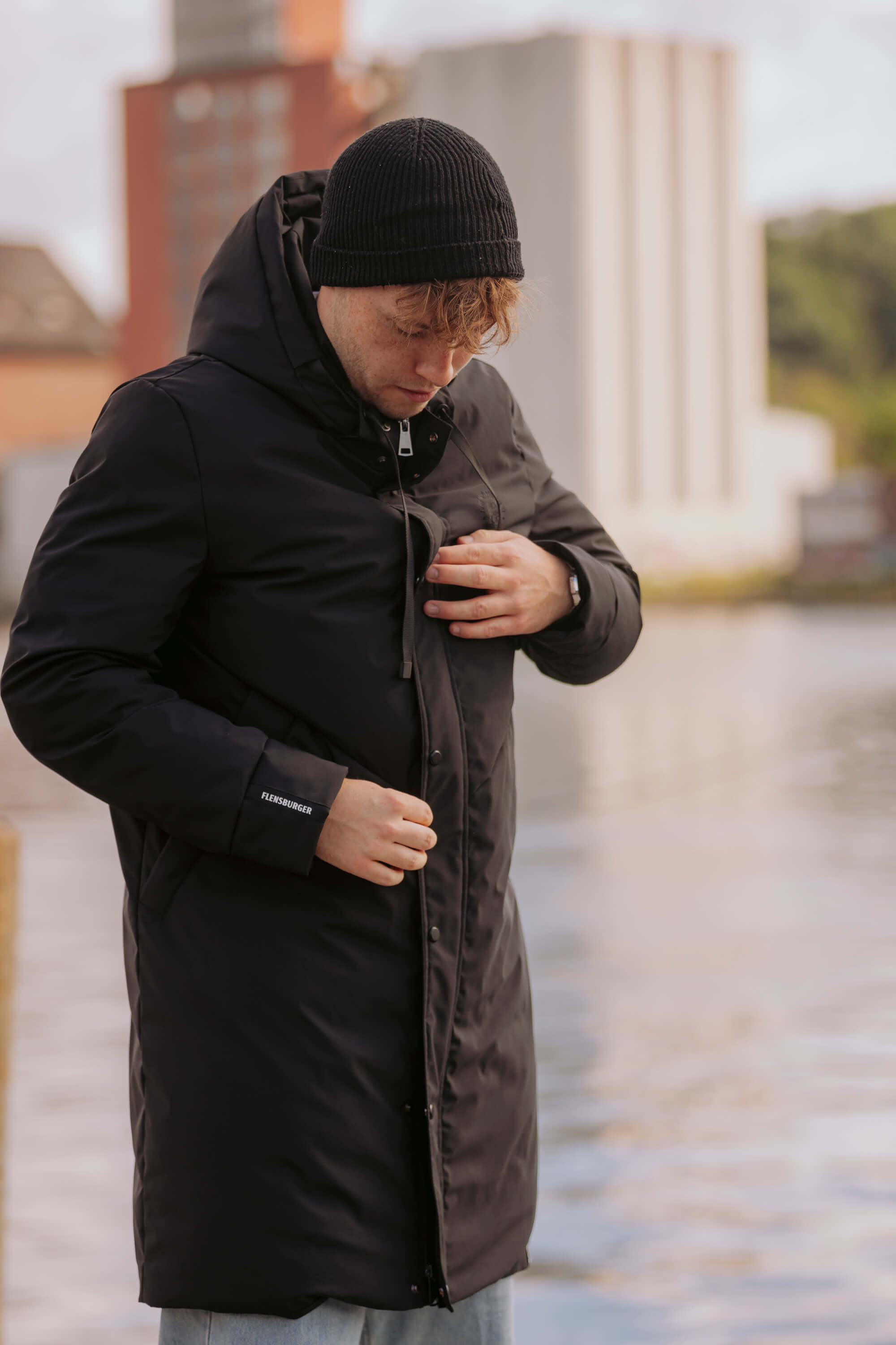 Ein junger Mann mit schwarzer Strickmütze trägt den schwarzen FLENS Winterparka "Ruben". Er schließt die Jacke mit beiden Händen. Am Ärmel ist das Logo "FLENSBURGER" sichtbar.