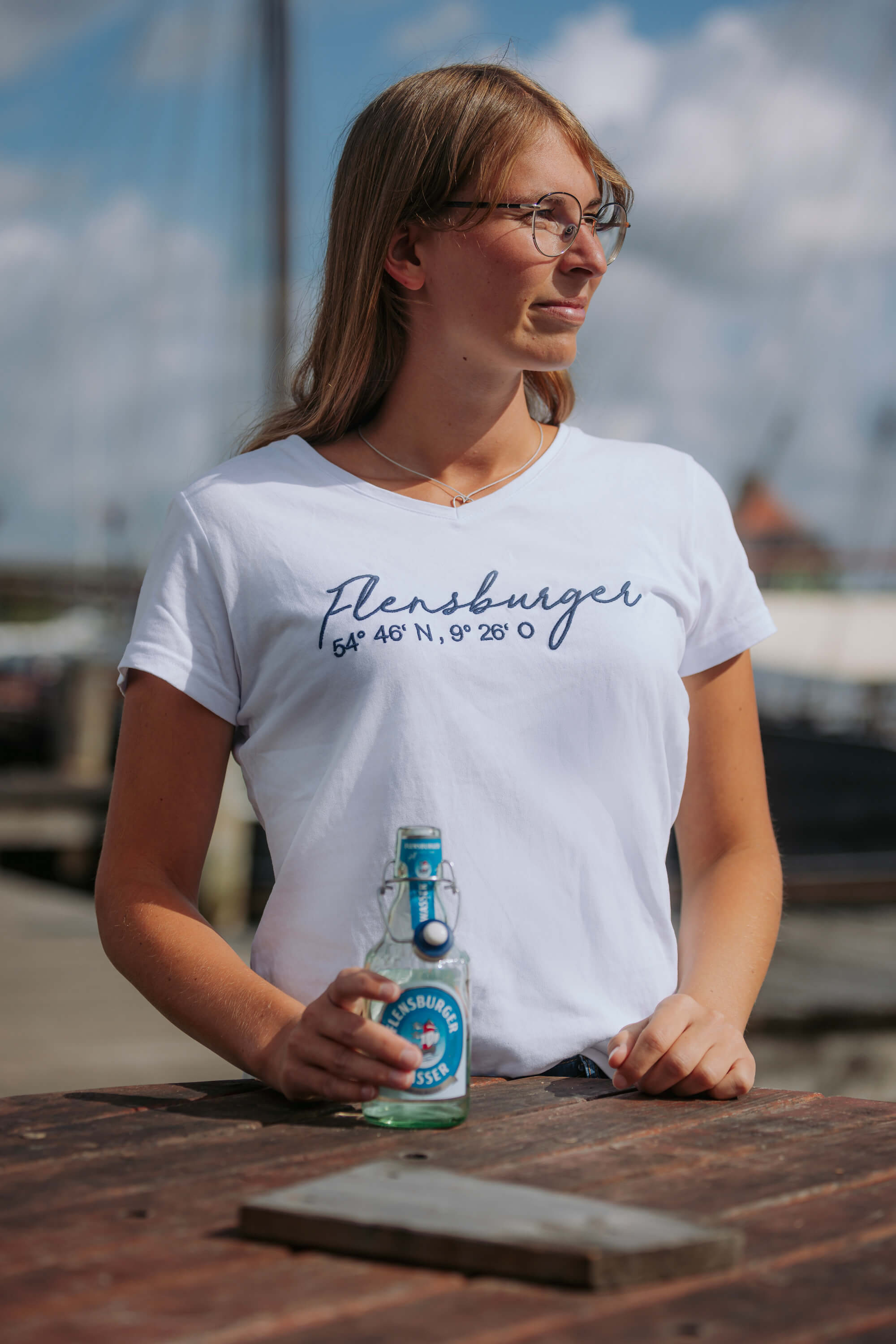 Frau trägt ein weißes T-Shirt mit dem geschwungenen Schriftzug "Flensburger 54° 46' N, 9° 26' O". Sie hält eine Flasche Flensburger Wasser und steht an einem Holztisch im Freien.