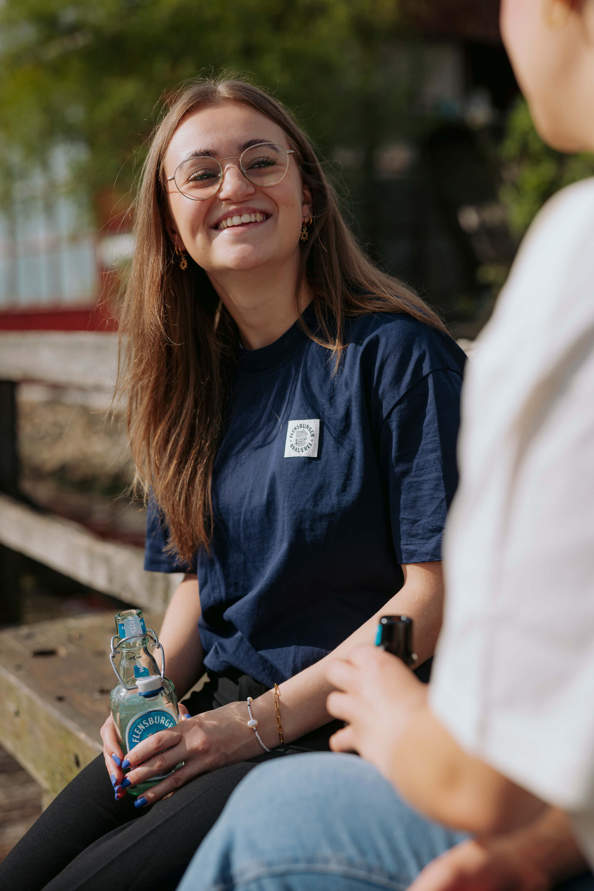 Eine junge Frau mit langen Haaren und Brille lächelt und hält eine Flasche Flensburger Wasser. Sie trägt ein dunkelblaues T-Shirt mit dem Logo der Flensburger Brauerei.