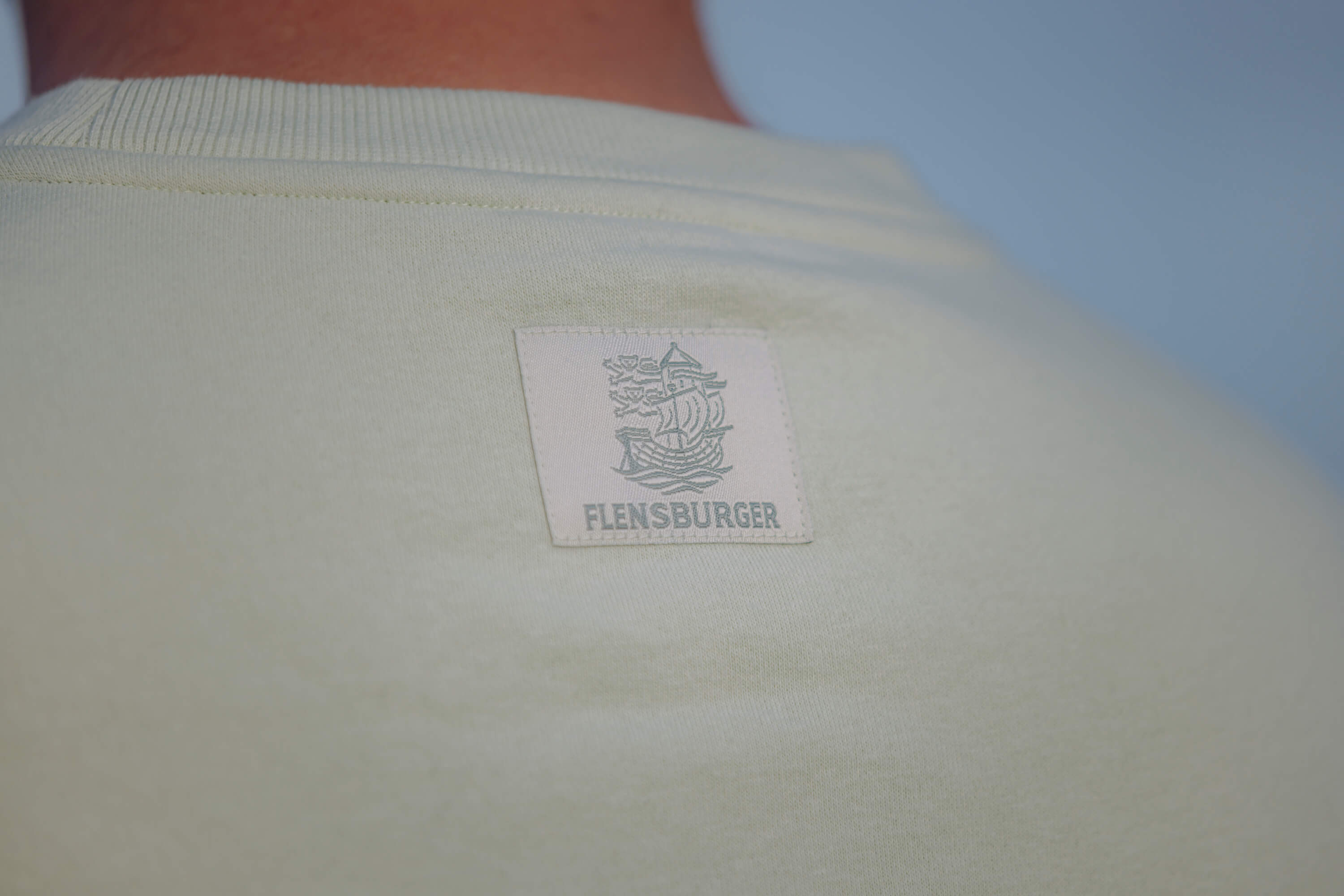 Detailaufnahme des weißen Sweaters "Sommerbrise" mit Aufnäher im Nachenbereicht, das ein stilisiertes Segelschiff und den Schriftzug "FLENSBURGER" zeigt.