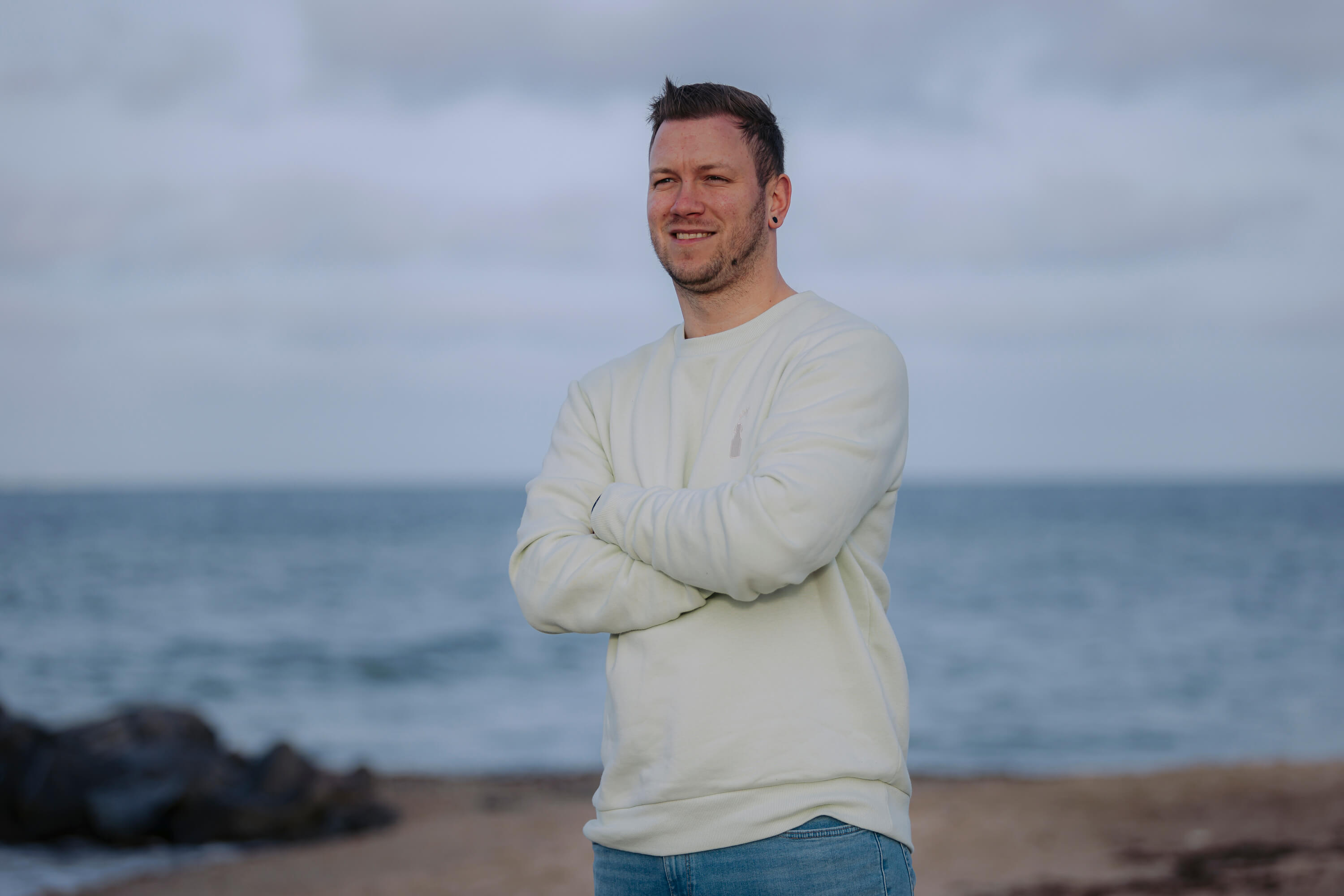 Junger Mann trägt den weißen Langarm-Sweater "Sommerbrise" mit Rundhalsausschnitt und steht mit verschränkten Armen am Strand vor dem Meer.
