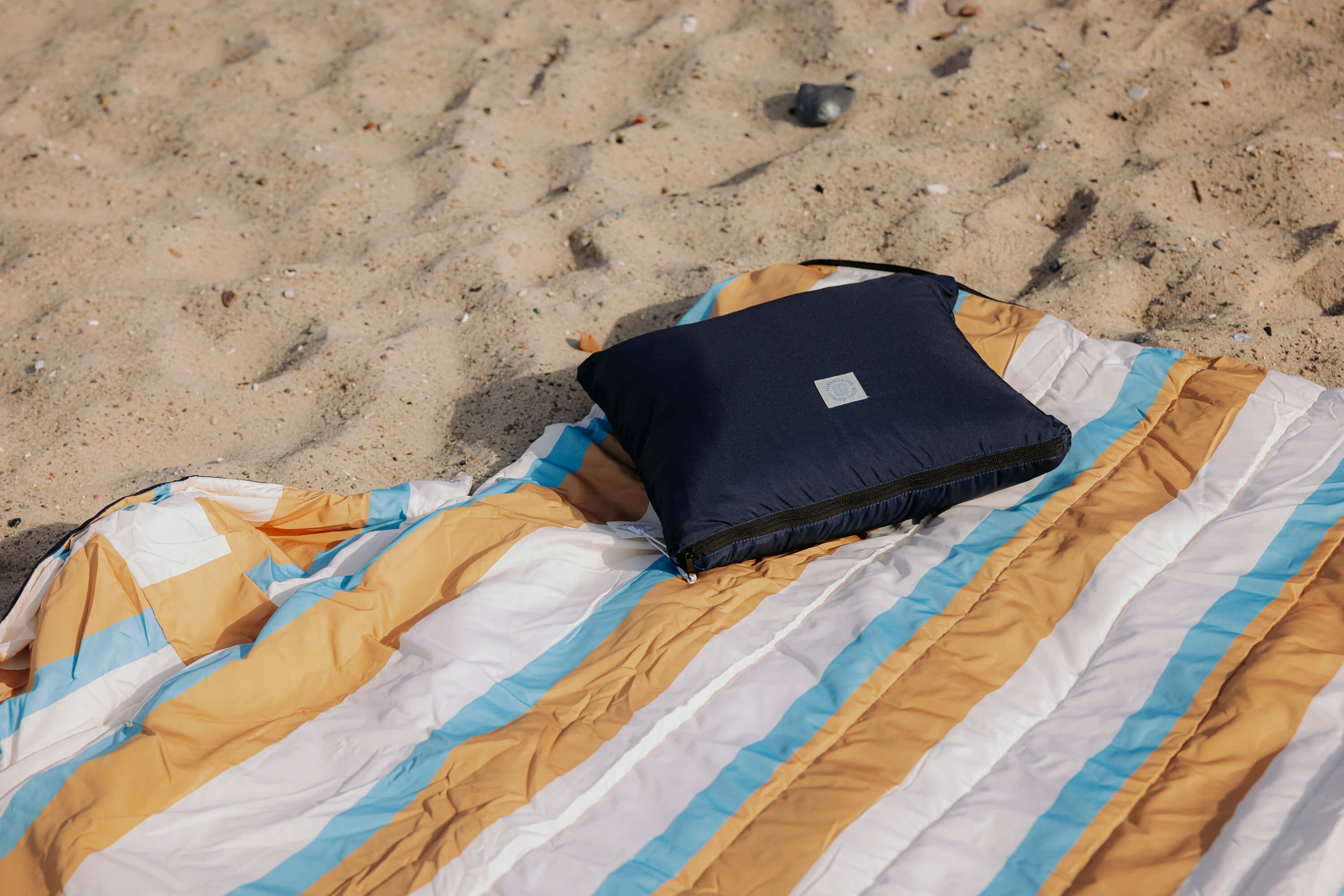 Eine gestreifte Outdoordecke in Beige, Braun, Weiß und Blau liegt auf sandigem Untergrund. Darauf liegt ein dunkelblaues, quadratisches Kissen mit einem weißen Aufnäher.