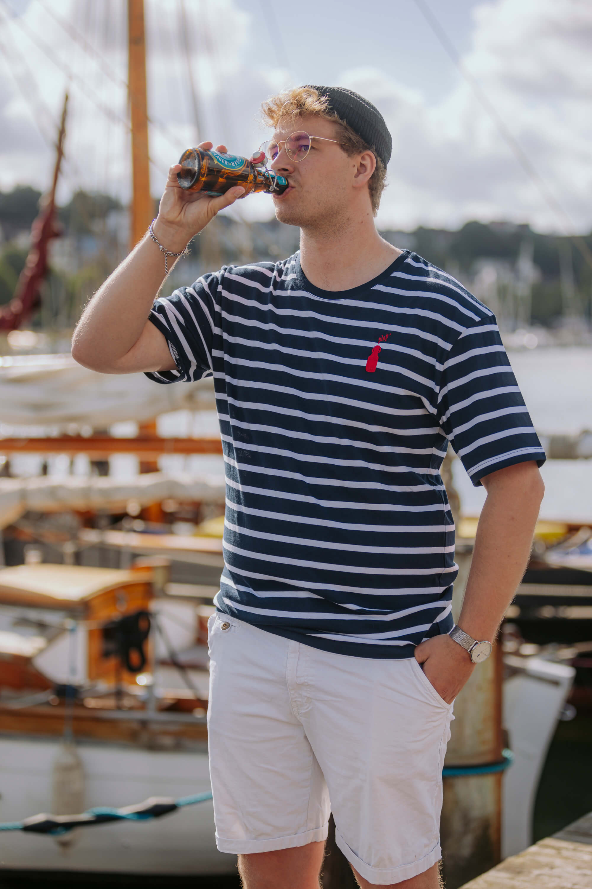 Mann mit schwarzer Mütze und Brille trinkt aus einerFlensburger Bügelflasche. Er trägt das blau-weiß gestreifte Plop’ & Stripes Shirt mit roter Flaschenstickerei und weiße Shorts. Im Hintergrund liegen Boote am Wasser.