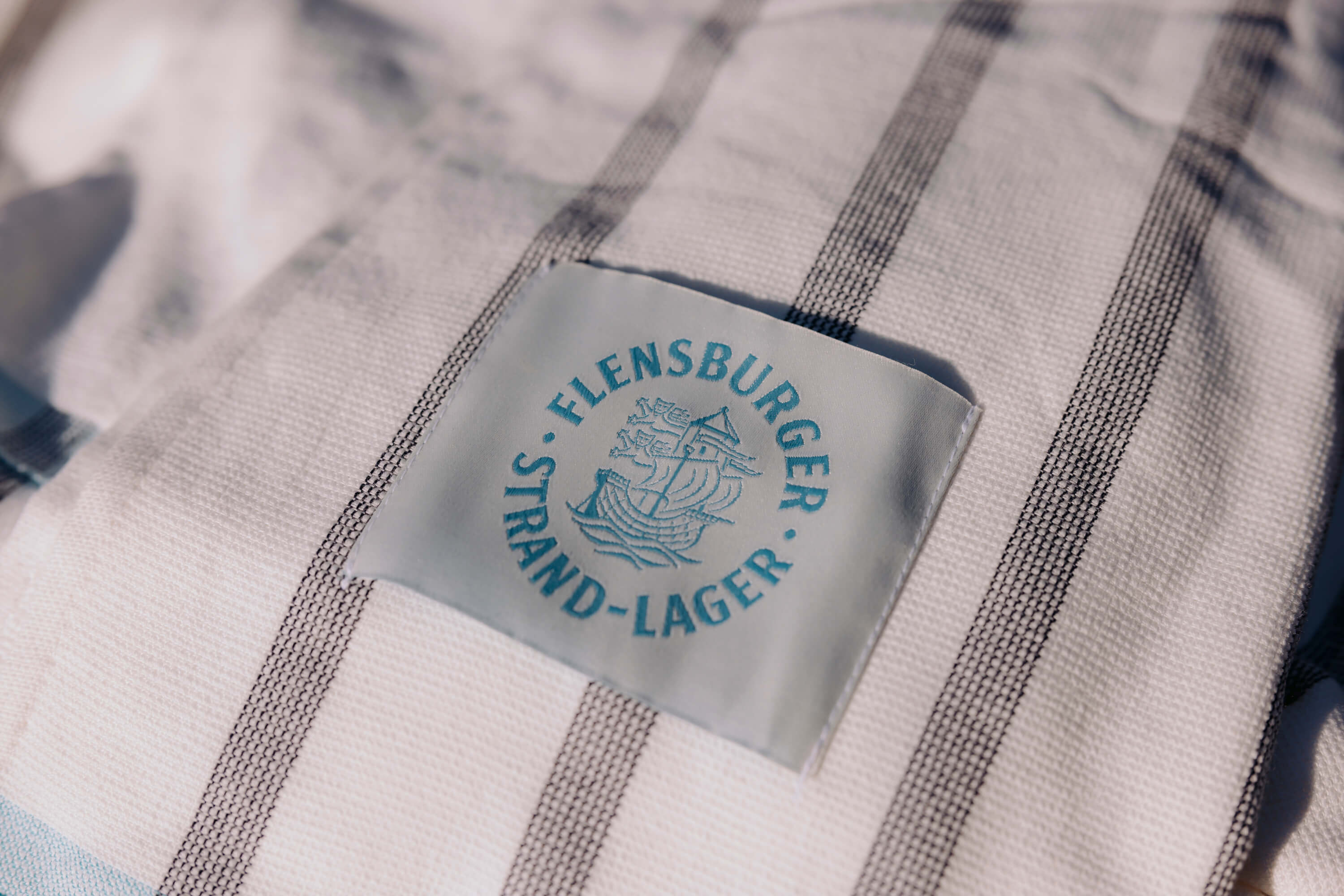 Detailansicht einer weiße Decke mit grauen Streifen zeigt ein eingenähtes Etikett mit Schriftzug "Flensburger Strand-Lager" und einem stilisierten Segelschiff in Blau.