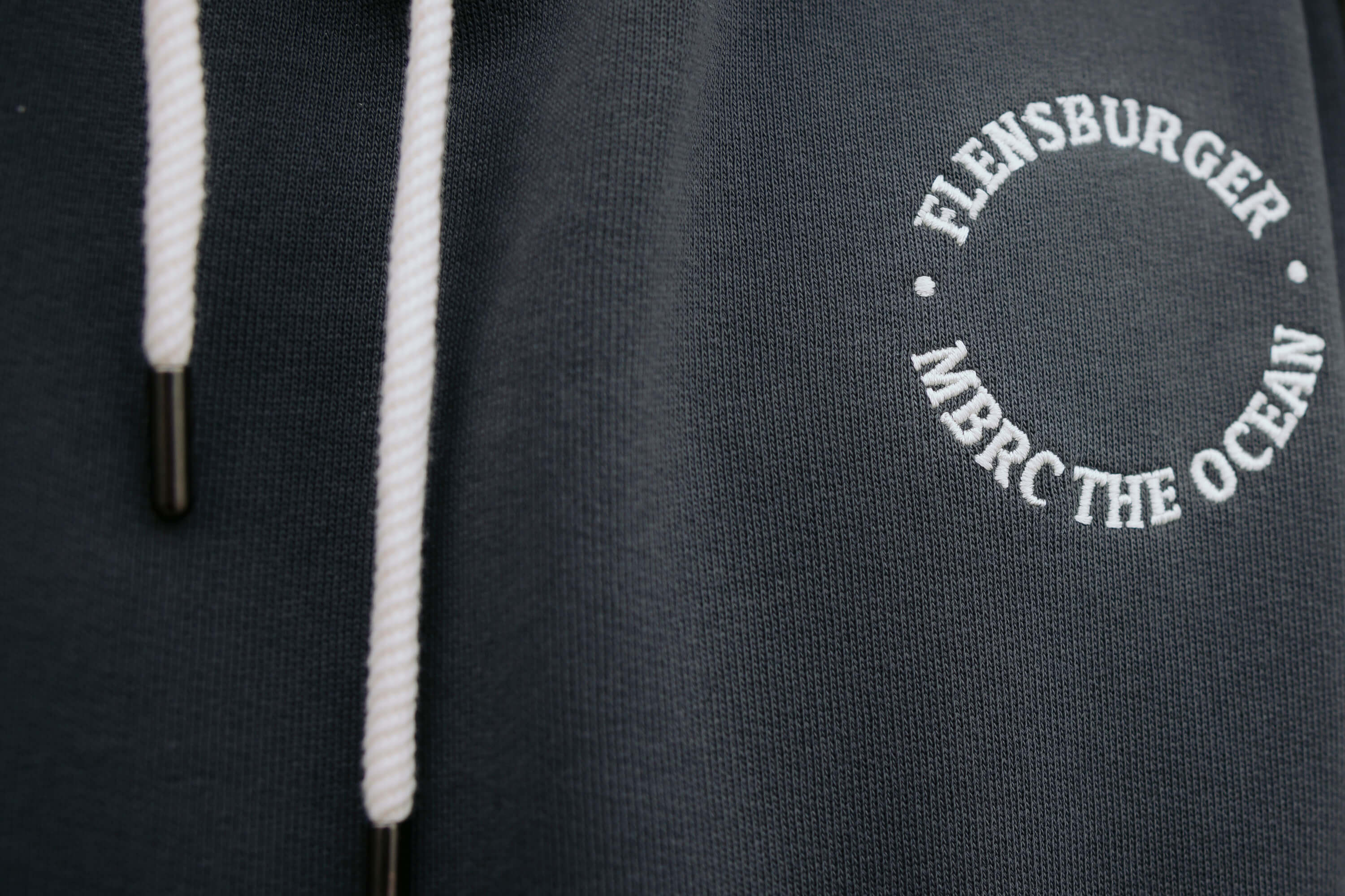 Detailaufnahme eines anthrazitfarbenen Hoodies mit weißen Kordeln. Auf der Brust ist weißer Text in Kreisform eingestickt: "FLENSBURGER . MBRC THE OCEAN .".