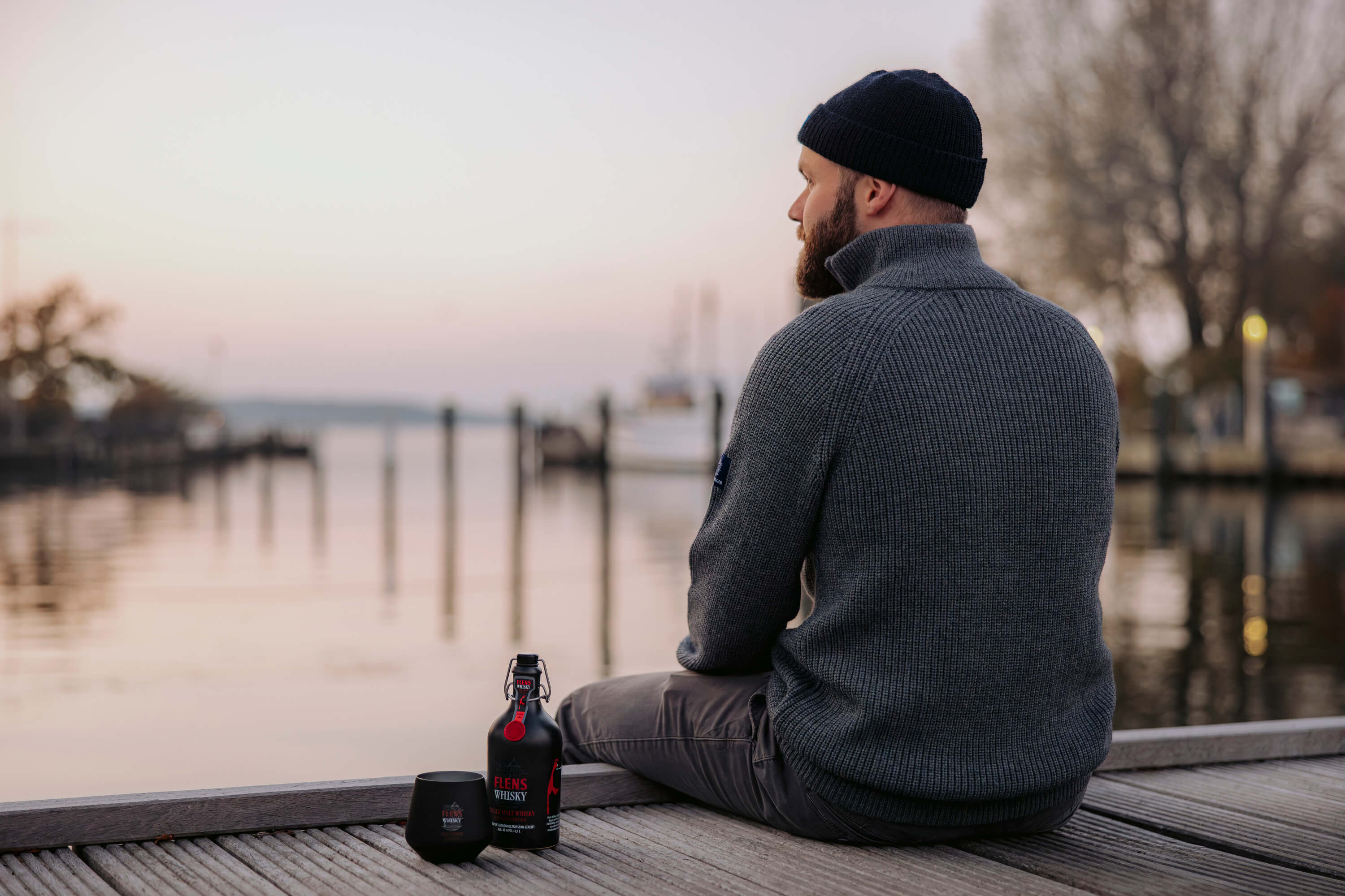 Mann mit anthrazitfarbenem Strickpullover und schwarzer Mütze sitzt auf einem Holzsteg und schaut auf das Wasser. Neben ihm steht eine Flasche FLENS Whisky und ein passendes schwarzes Glas.