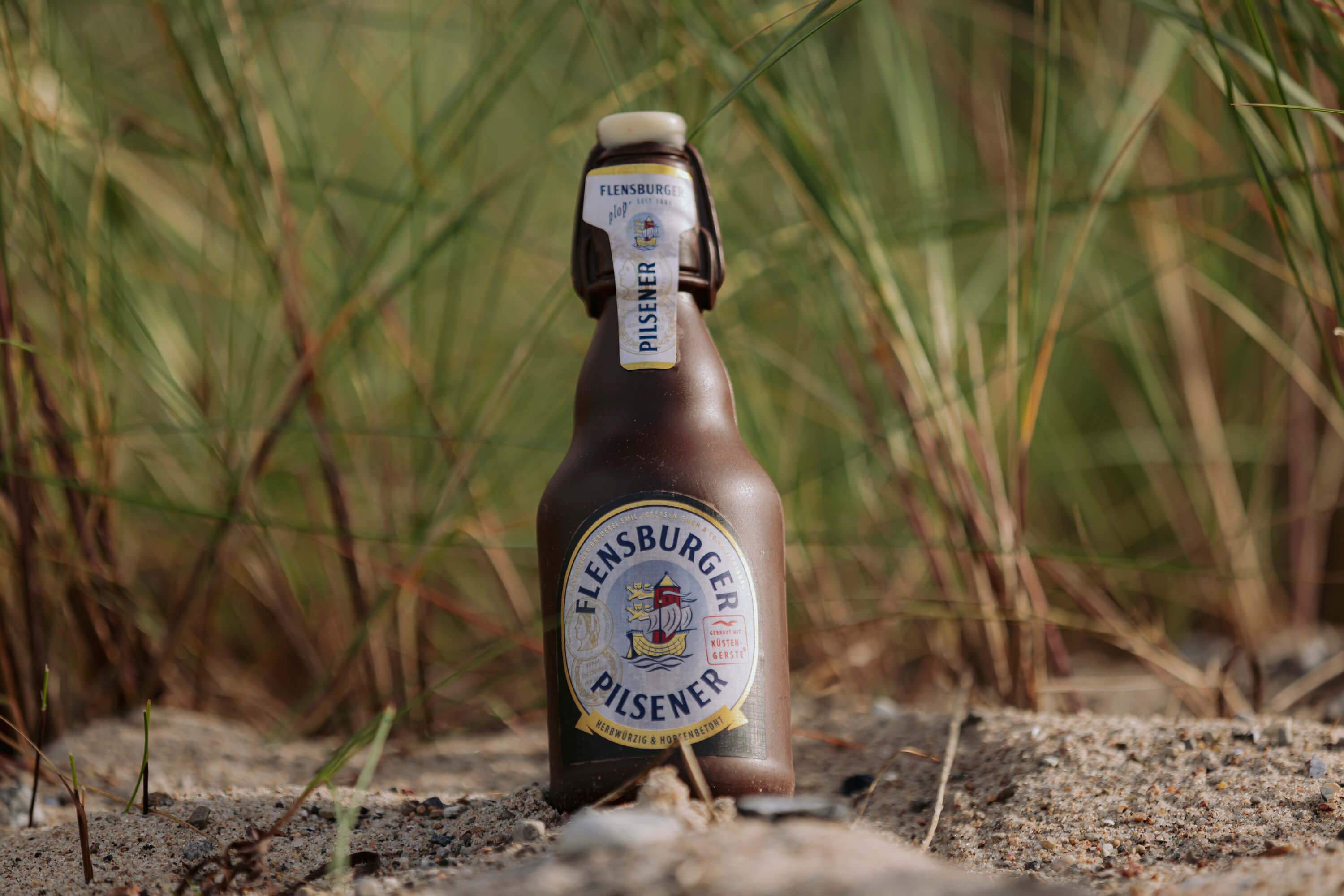 Flensburger Pilsener Schokoladen-Flasche in 0,3 l Größe steht auf Sand vor grünen Halmen. Das Etikett zeigt das Flensburger Logo und die Wörter „herbwürzig & hopfenbetont“.