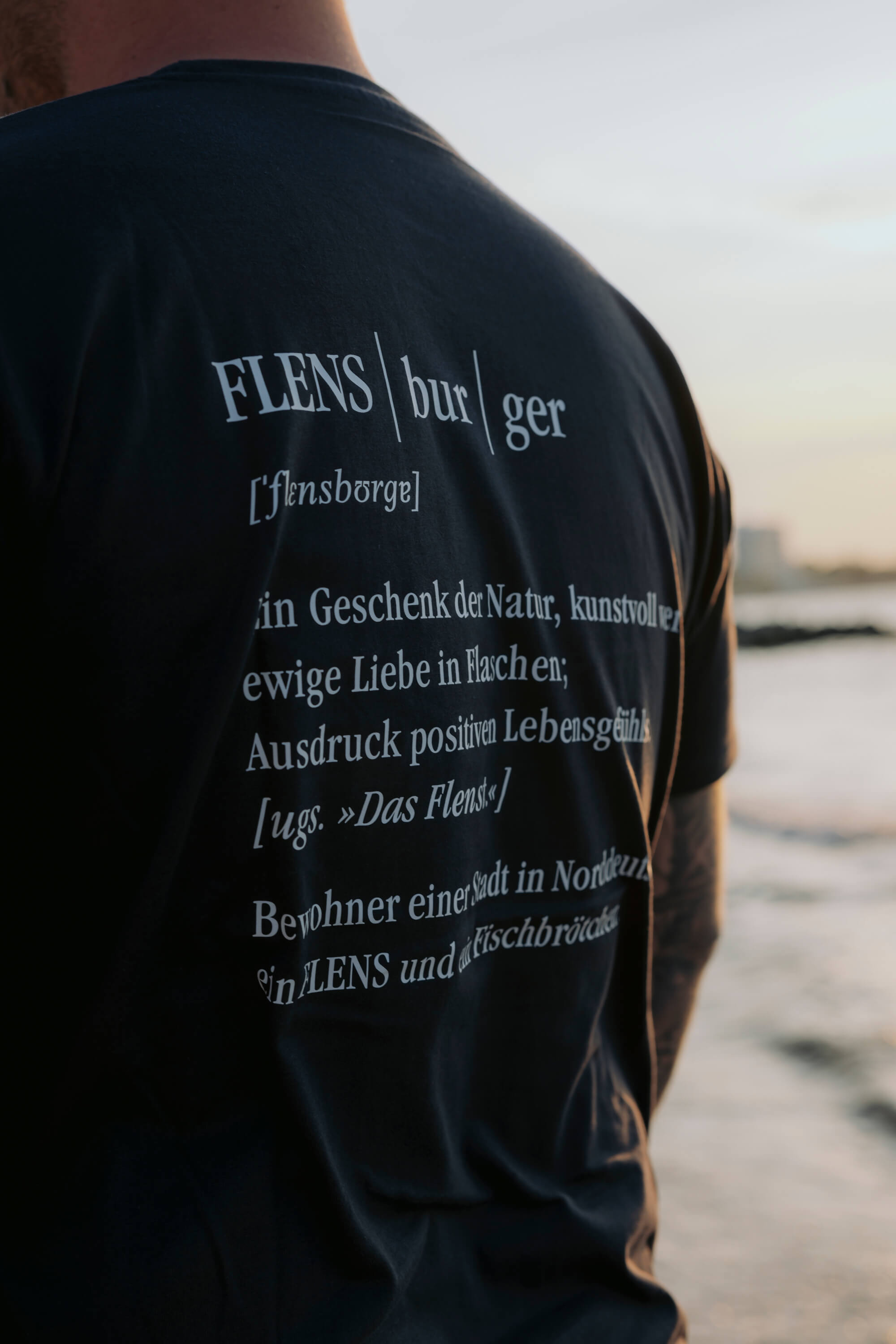Person trägt ein schwarzes T-Shirt, mit weißem Text auf dem Rücken. Darauf seht "FLENS | bur | ger", phonematische Aussprache [ˈflɛnsbɔʁɡɐ] und Definitionen, teilweise kursiv gedruckt. Im Hintergrund ist ein Strand zu sehen.