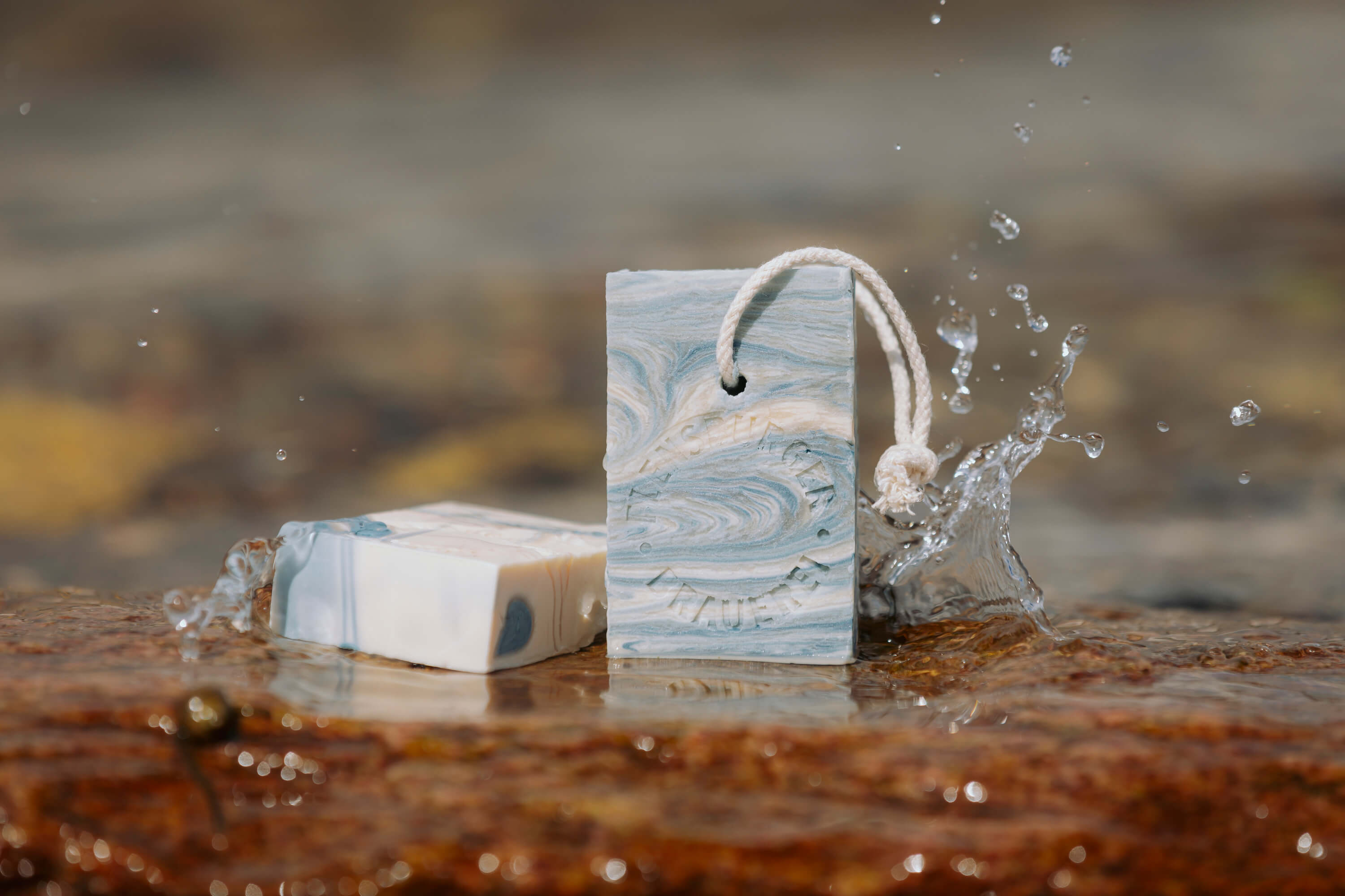 Zwei Stücke feste Duschseife in  Blau-Weiß mit Marmorierung liegen auf nassem Stein. Eines steht aufrecht mit einer Schlaufe, das andere liegt flach. Wasser spritzt daneben.
