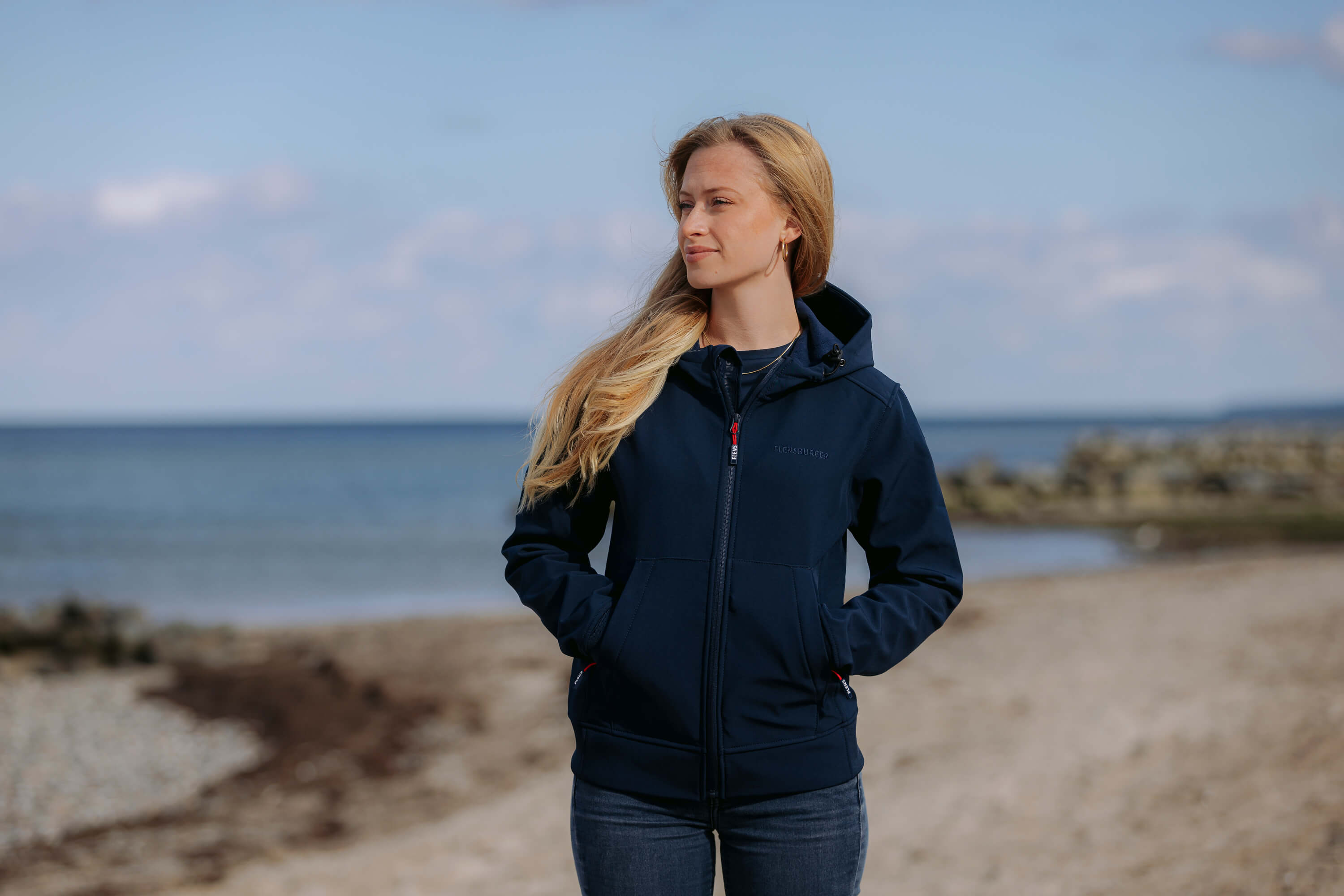 Eine Frau mit blonden langen Haaren trägt eine navyblaue Softshelljacke mit Kapuze und Reißverschluss. Sie steht am Strand mit dem Rücken zum Meer. Ihre Hände stecken in den Taschen der Jacke.