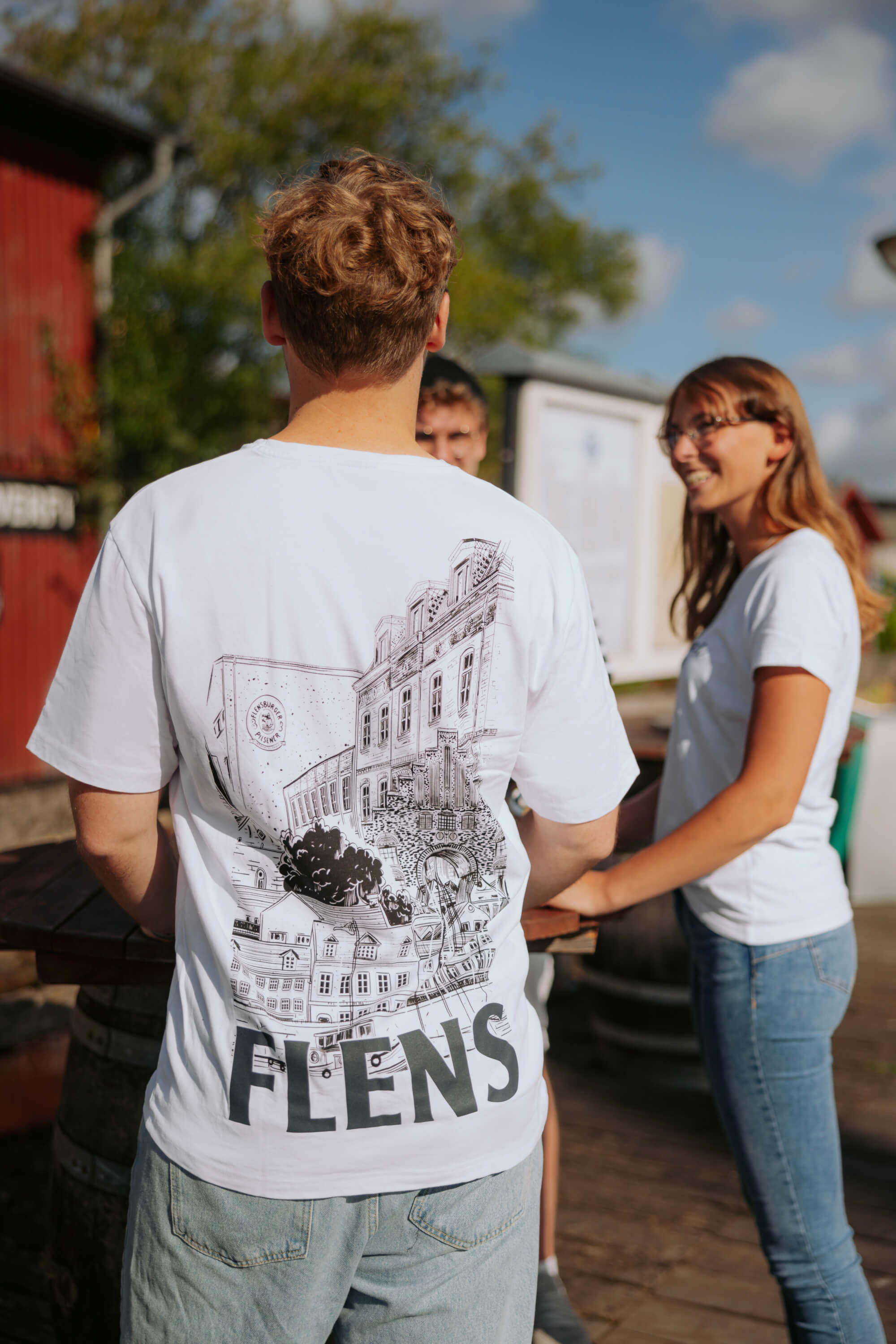 Ein Mann trägt ein weißes Oversize-T-Shirt mit einem schwarzen, detaillierten Stadtmotiv und dem Schriftzug „FLENS“ auf dem Rücken. Zwei Personen stehen ihm gegenüber.