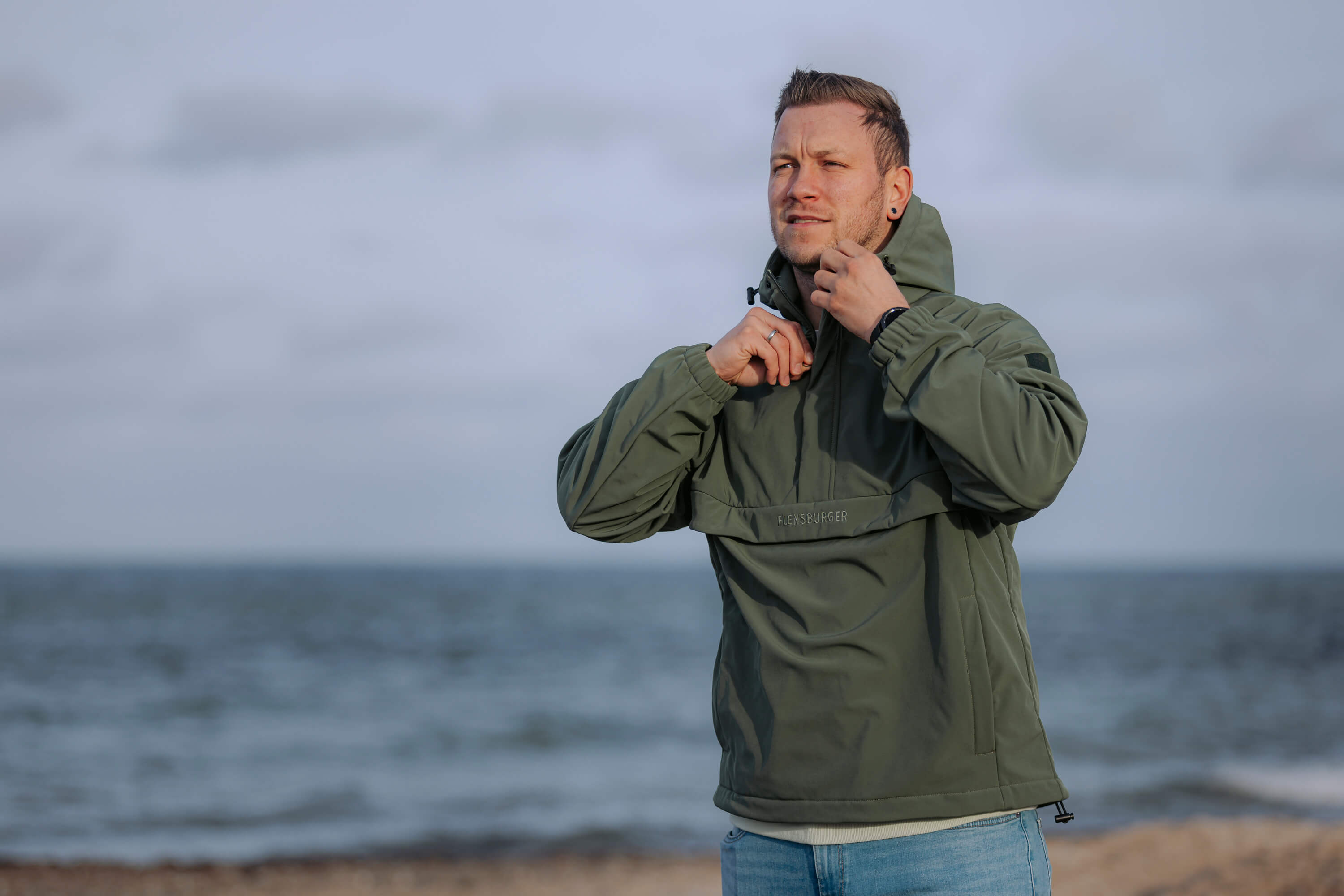 Mann steht am Strand und zieht den Reißverschluss eines olivgrünen, wetterfesten Windbreakers der Marke Flensburger hoch.