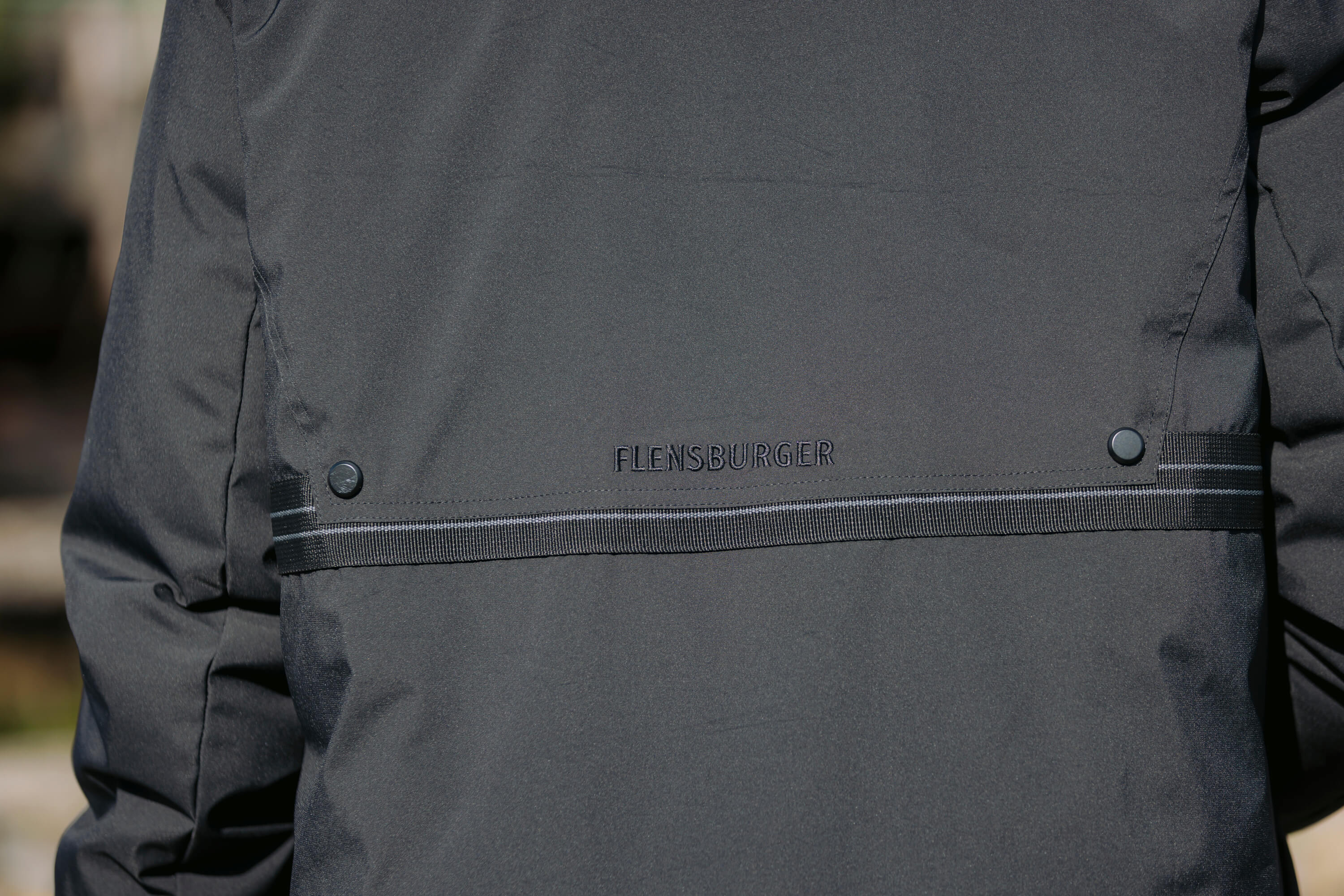 Der schwarze Winterparka zeigt auf dem Rücken den Schriftzug „FLENSBURGER“ in schwarzer Stickerei. Der Parka hat zwei Druckknöpfe und einen horizontalen  Streifen.