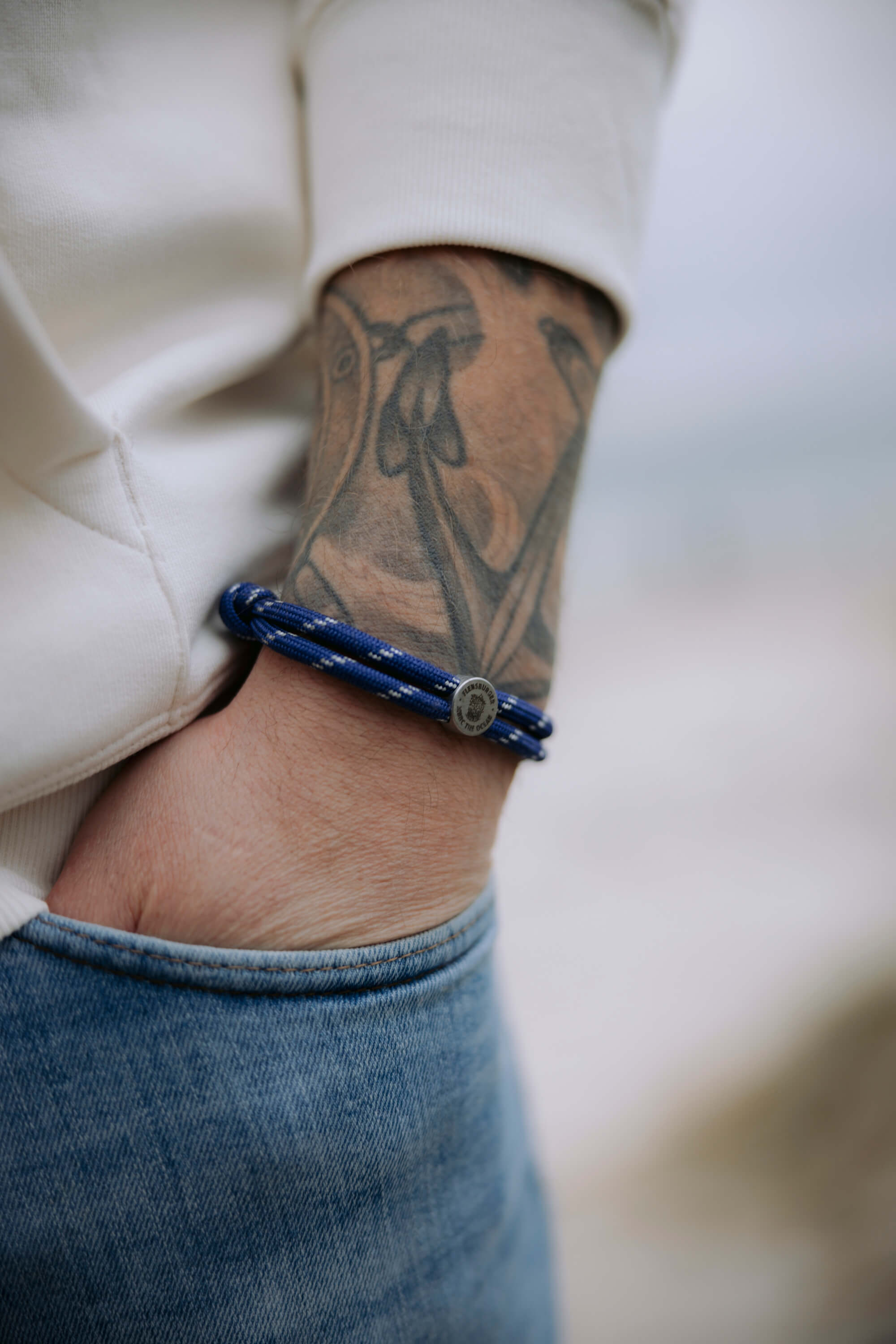 Ein Arm mit einer schwarzen Tätowierung trägt ein blau-weißes Armband aus Garn aus 100% rPET. Die Hand steckt in der Tasche einer blauen Jeans.