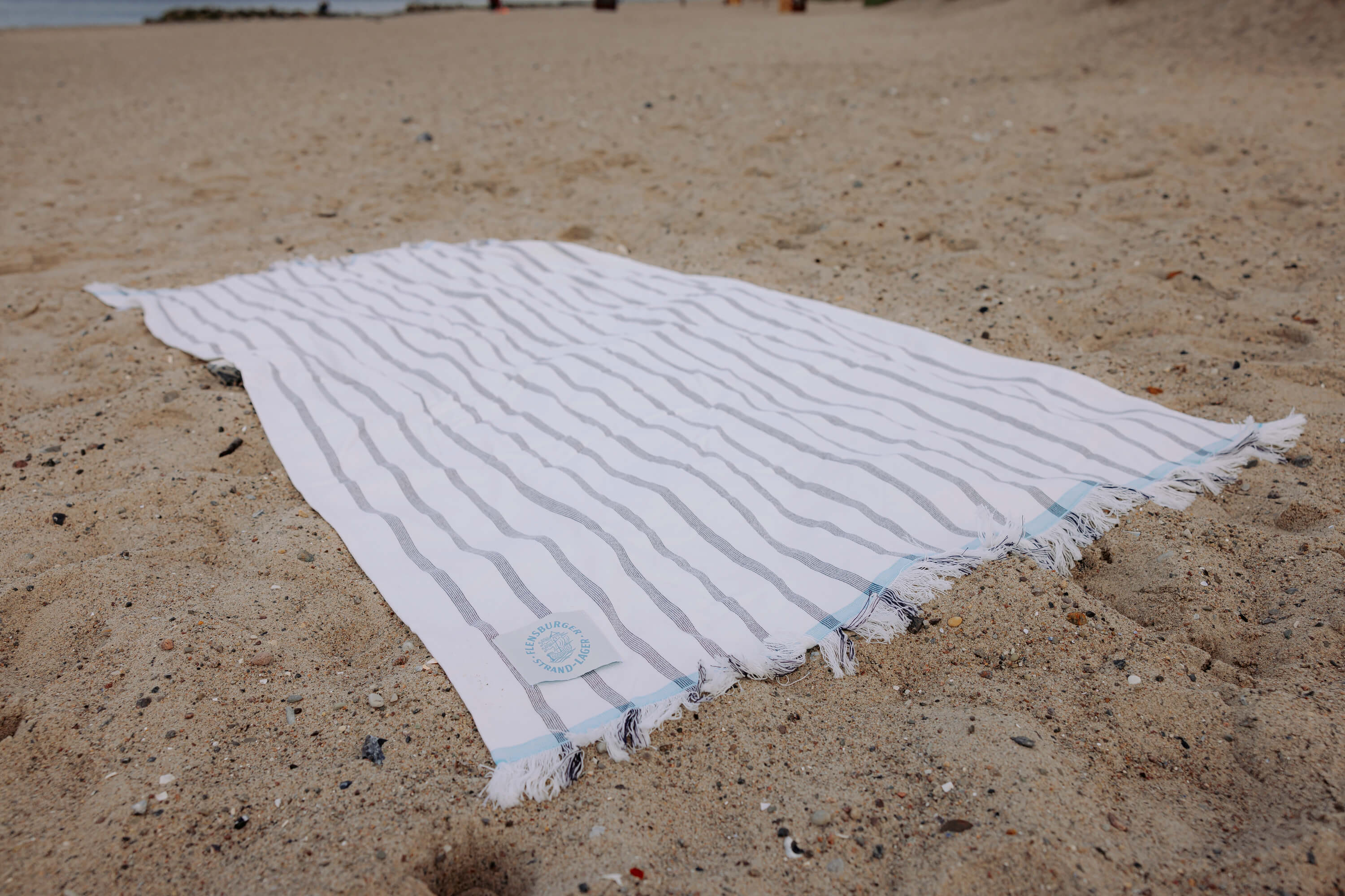 Weiße Stranddecke mit grauen Streifen liegt auf Sand. Am unteren Rand ist ein weißer Aufnäher mit runder aufgedrucktem blauem Text "Flensburger Strand Lager".