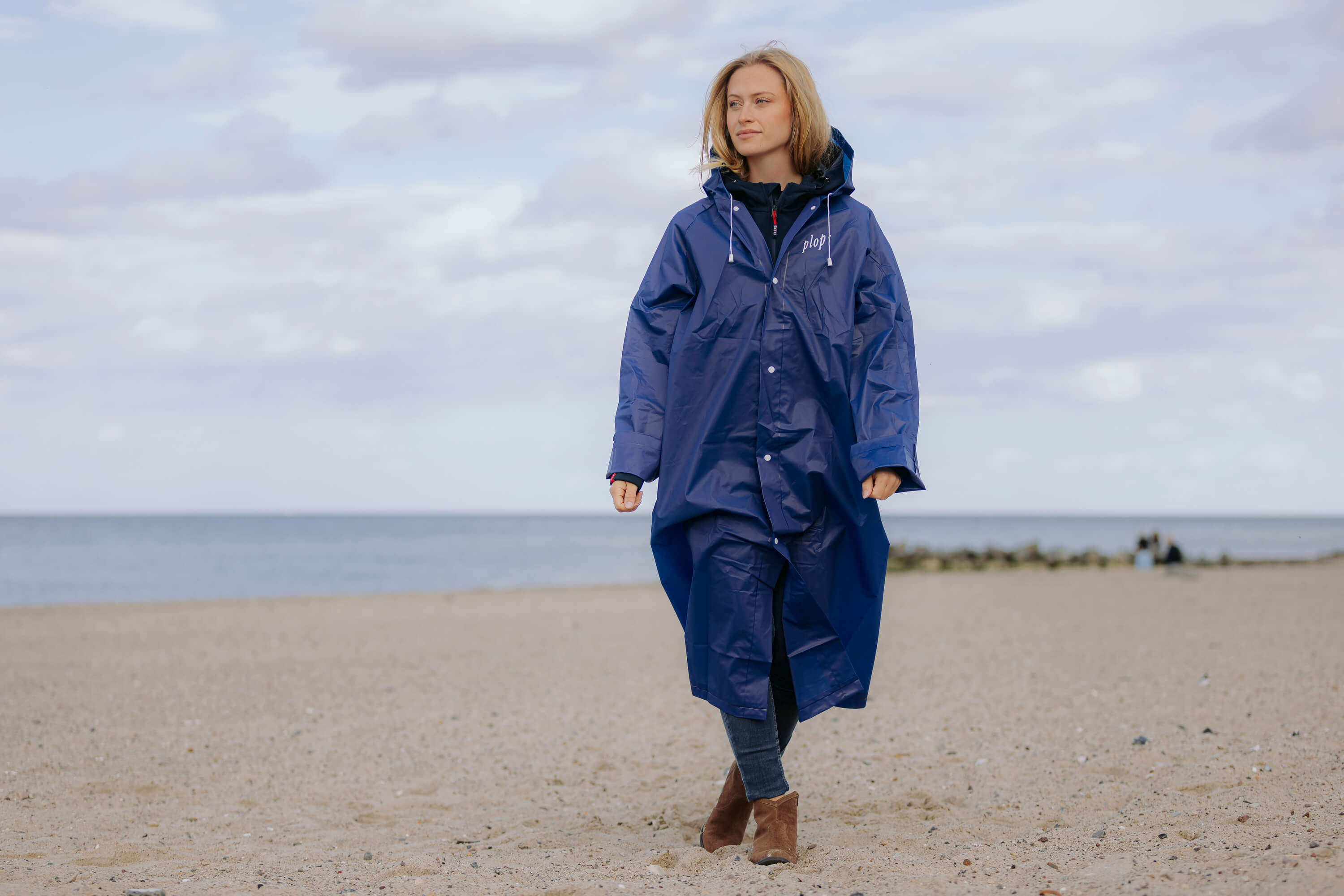 Eine Frau trägt den dunkelblauen FLENS Regenponcho mit Kapuze. Sie steht am Strand vor dem Meer. Der Poncho hat vorne rechts die Aufschrift „plop'“ in Weiß.