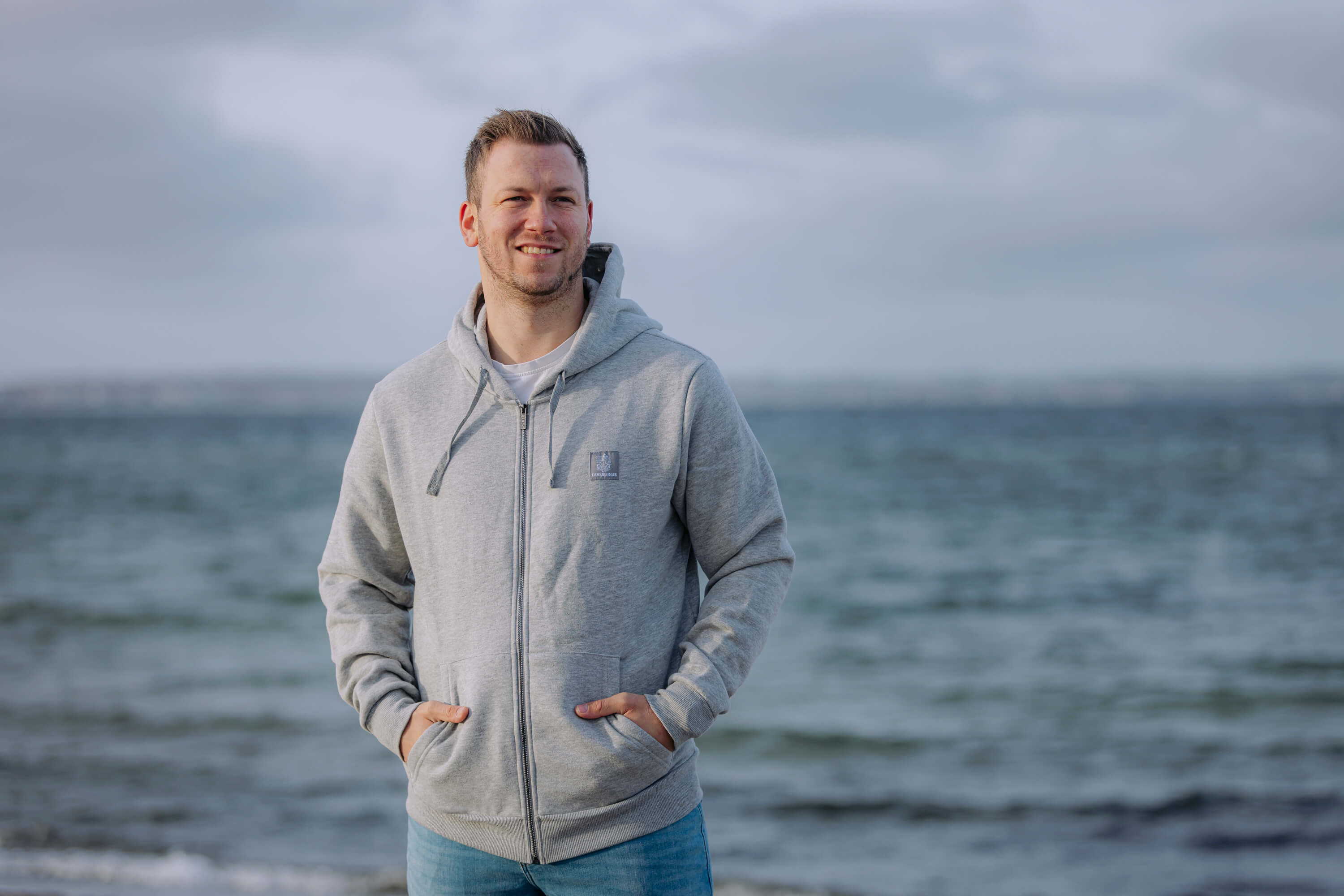 Mann trägt eine hellgraue Sweatshirtjacke mit Kapuze und Reißverschluss. Er steht mit den Händen in den jackentaschen am Meer. Der Himmel ist bewölkt.