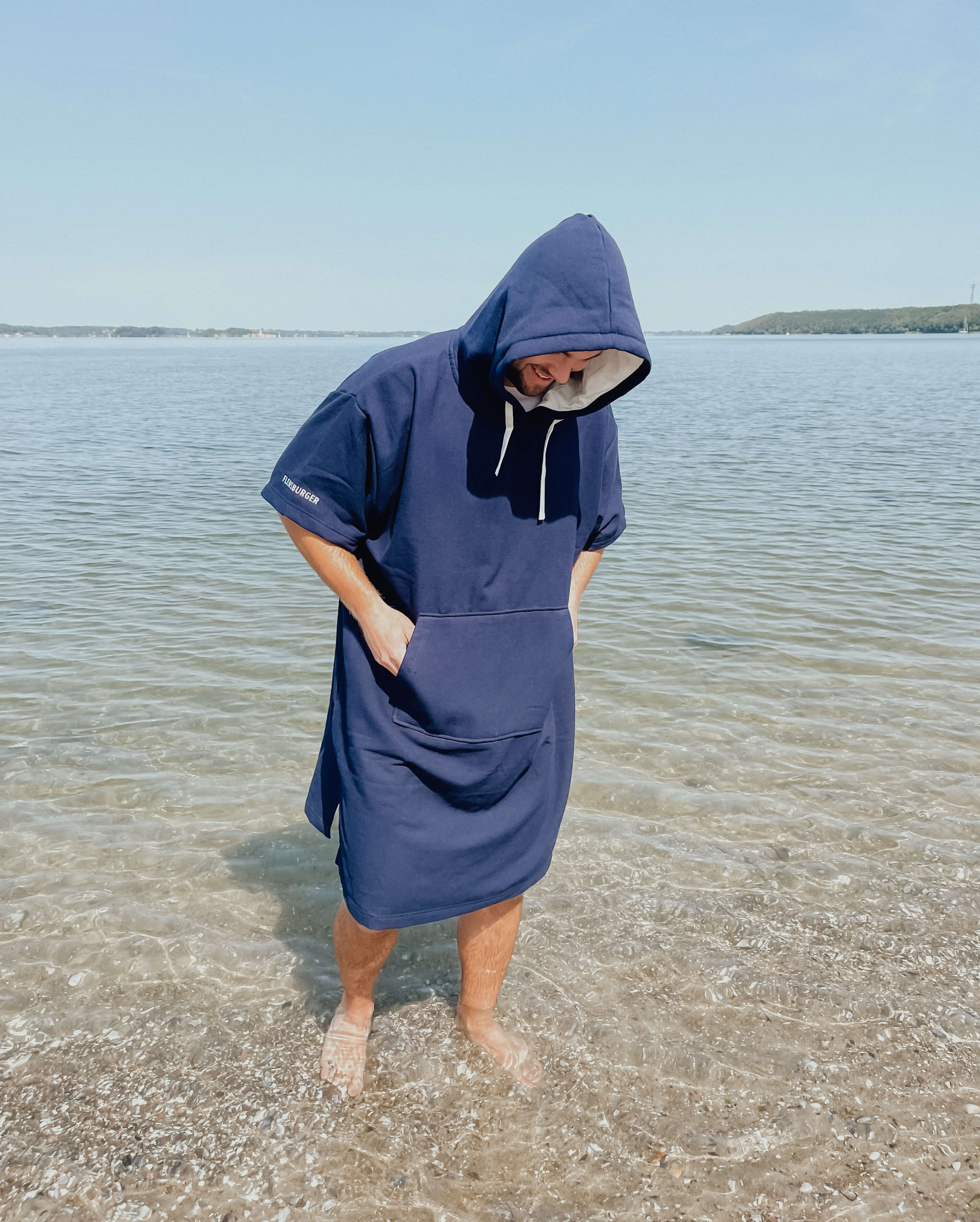 Ein Mann trägt einen blauen Kapuzen-Strandponcho aus Frottee und steht barfuß im flachen Wasser an einem Strand. Sie schaut nach unten ins Wasser und hat die Hände in den Ponchotaschen gesteckt.