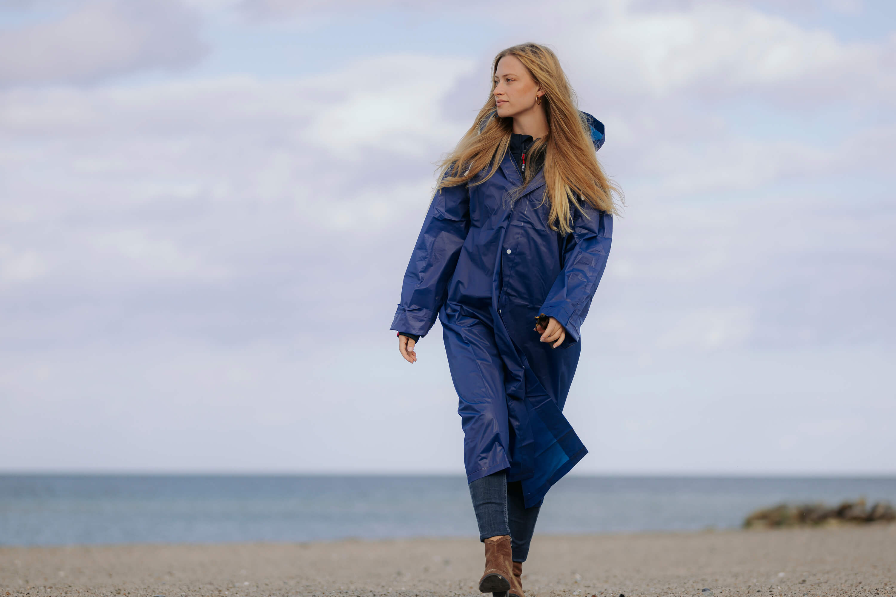 Eine Frau läuft am Strand und trägt einen langen, blauen Regenponcho mit Kapuze.