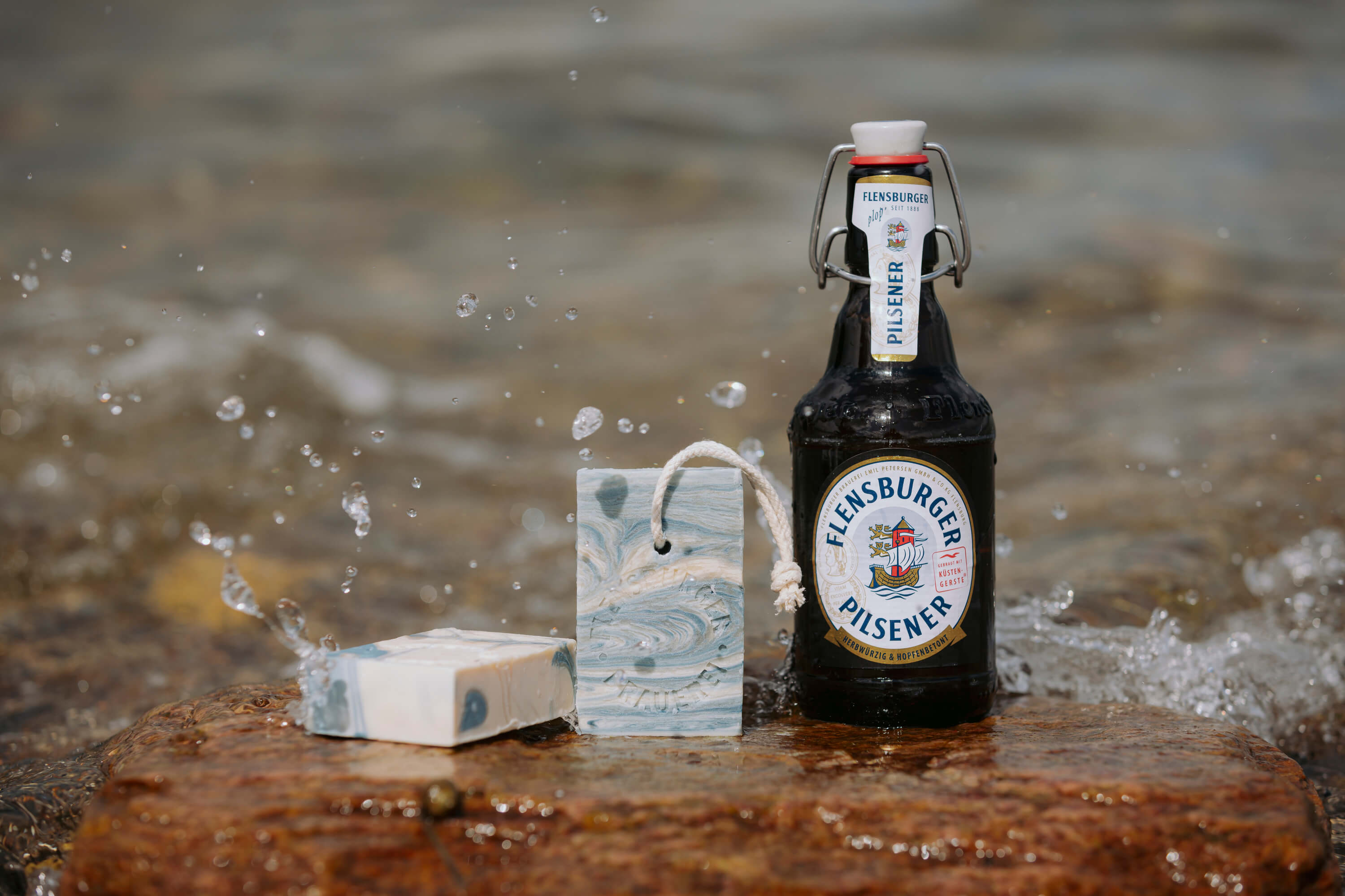 Eine Flasche Flensburger Pilsener steht auf einem Stein, daneben liegen zwei blau-weiß marmorierte Seifenstücke, um die das Wasser spritzt.