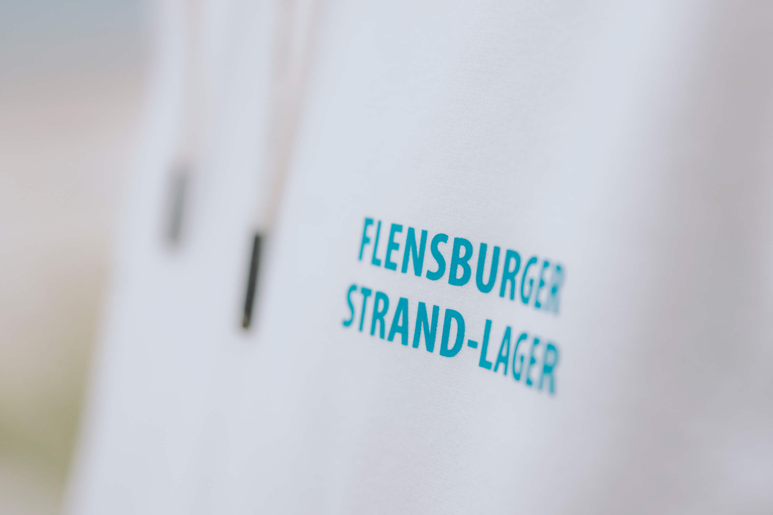 Detailaufnahme der Aufschrift "FLENSBURGER STRAND-LAGER" in türkisfarbener Schrift auf der Vorderseite eines beigen Hoodies.