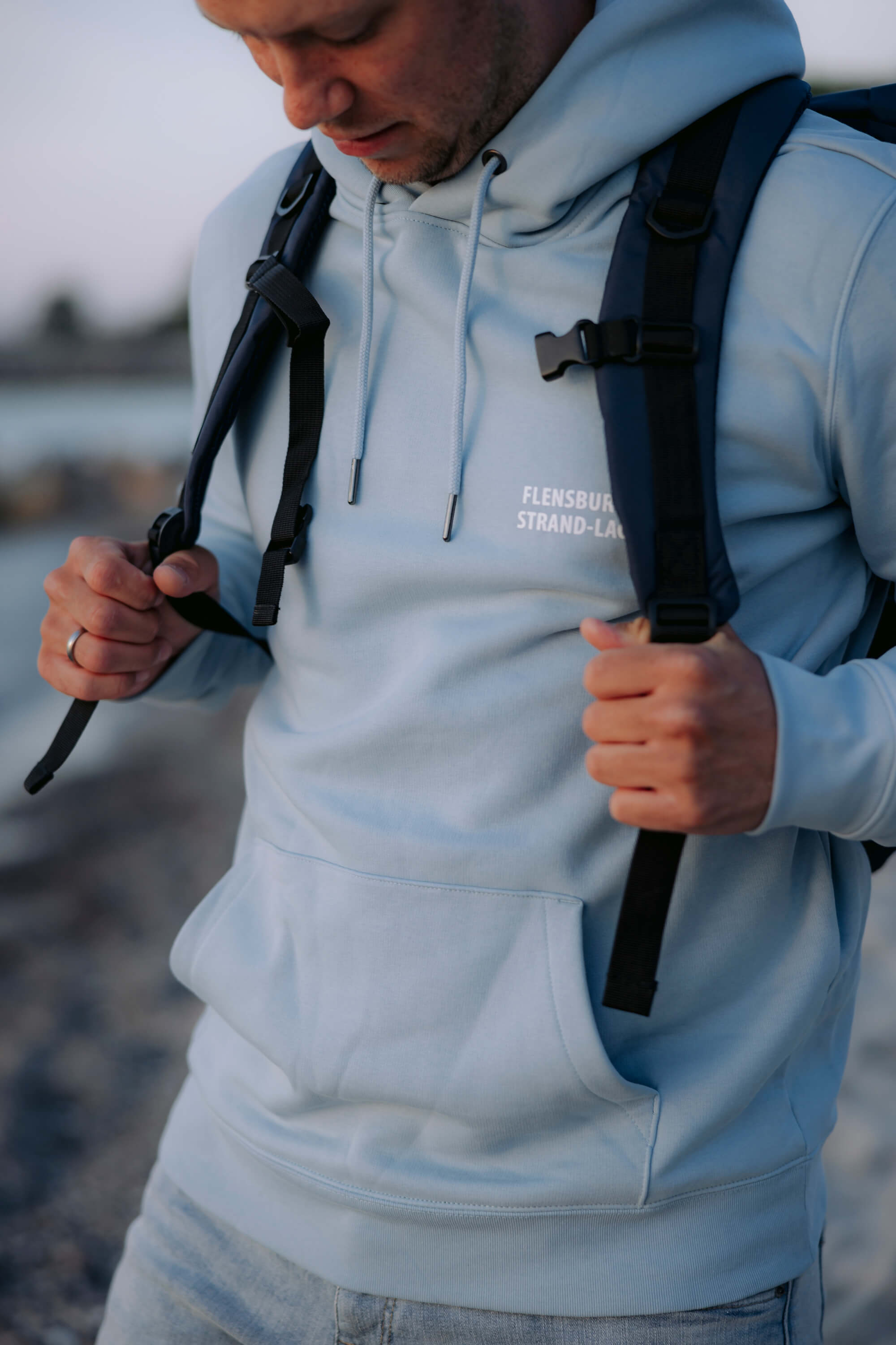 Mann trägt hellblauen Hoodie mit der weißen Aufschrift "Flensburg Strand-Lager". Er hält die Träger eines dunkelblauen Rucksacks namens "Nordlicht Explorer" fest.