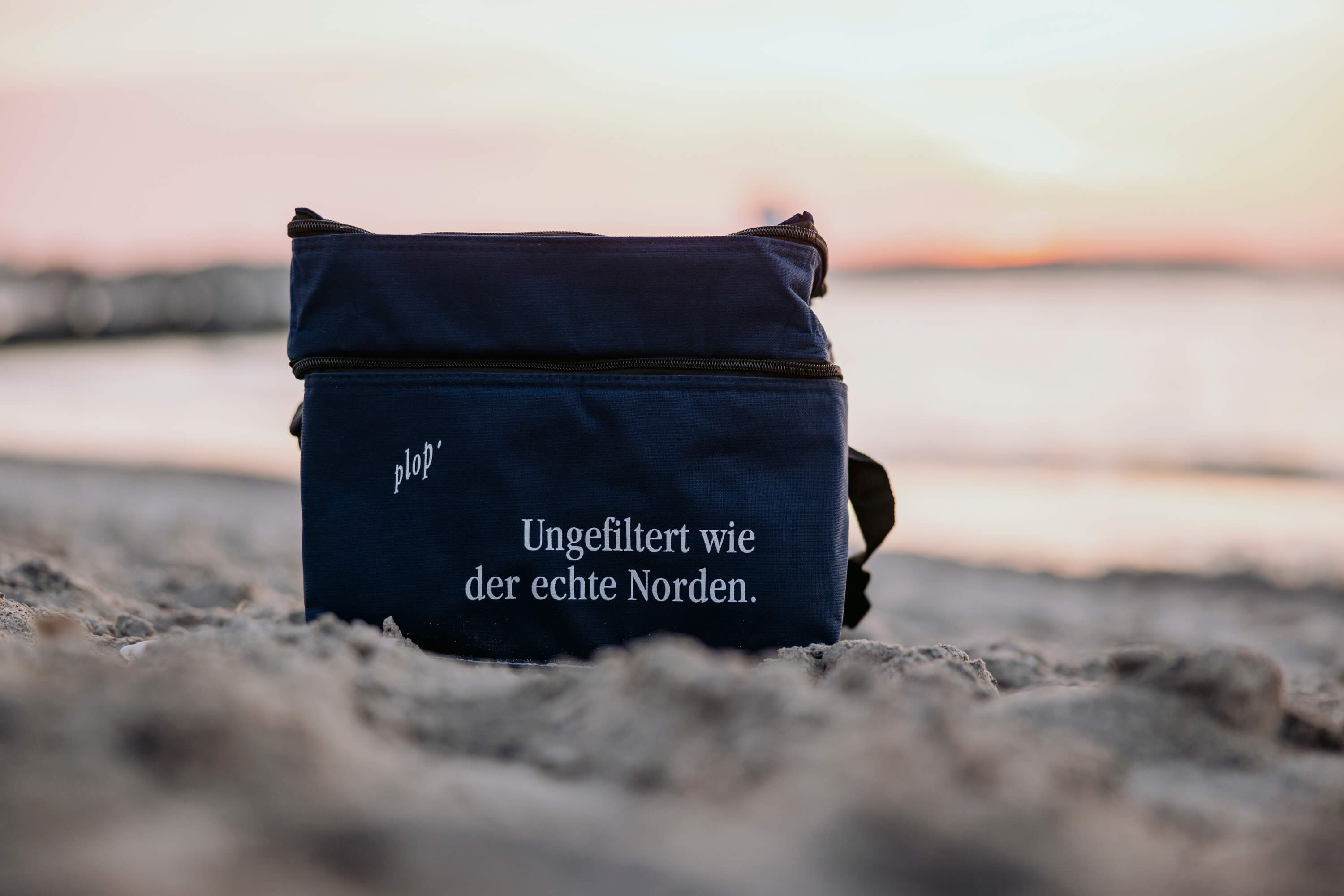 Kühltasche in Navyblau mit dem Text "Ungefiltet wie der echte Norden" steht auf einem Sandstrand. Die Tasche ist offen, während die Wellen im Hintergrund sanft an den Strand rollen.