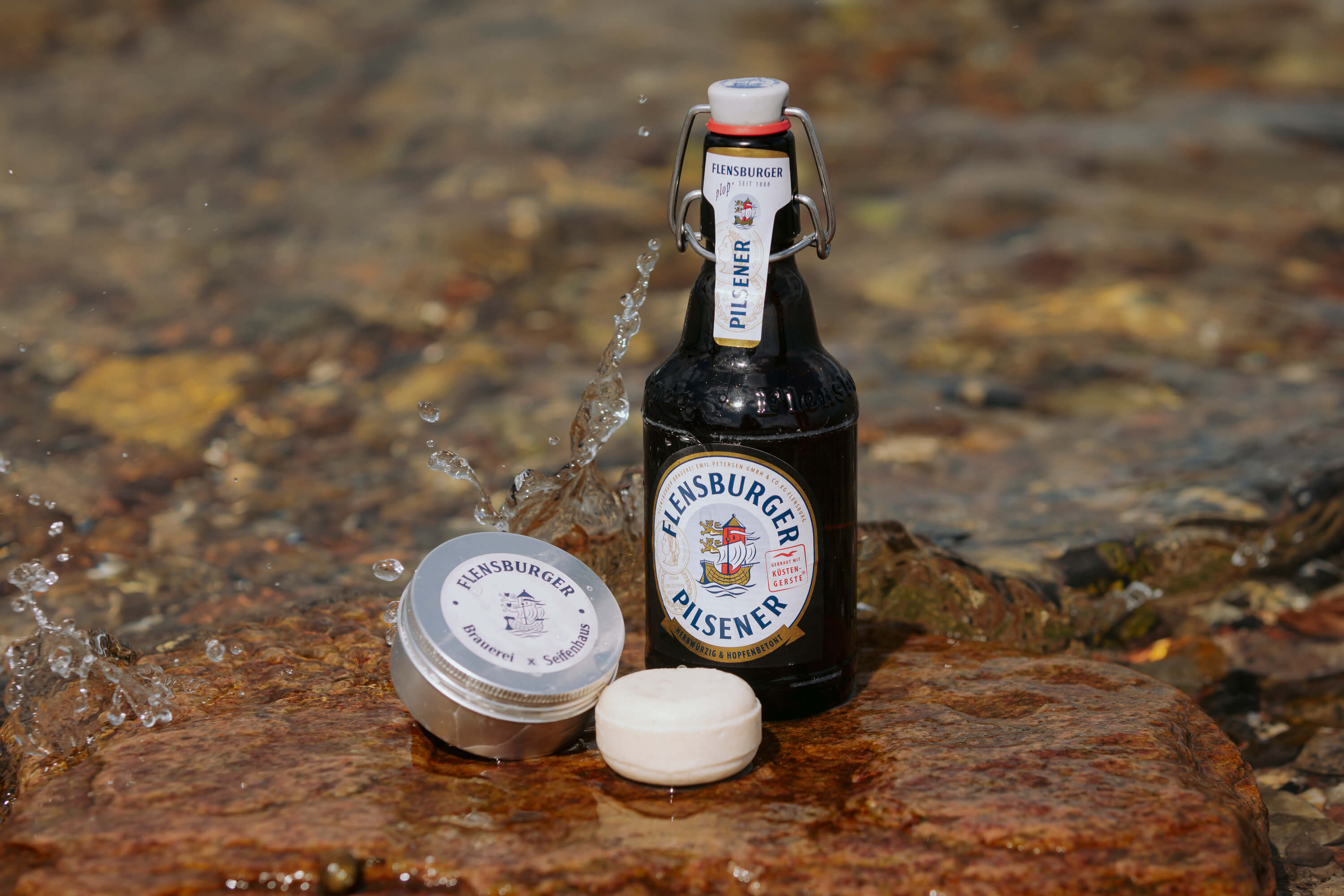 Flensburger Pilsener Flasche steht auf einem nassen Stein. Daneben liegen ein rundes, weißes festes Shampoo und eine Dose mit der Aufschrift "Flensburger Brauerei x Seifenhaus".