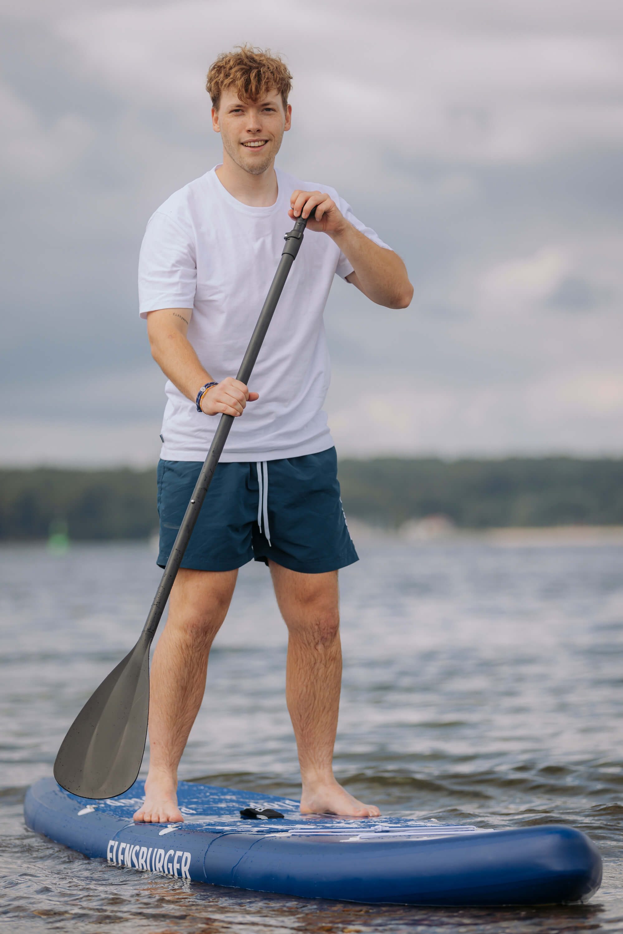 Junger Mann steht auf einem blauen SUP-Board mit der Aufschrift „FLENSBURGER“. Er hält im Stehen ein Paddel und trägt ein weißes T-Shirt sowie blaue Shorts.