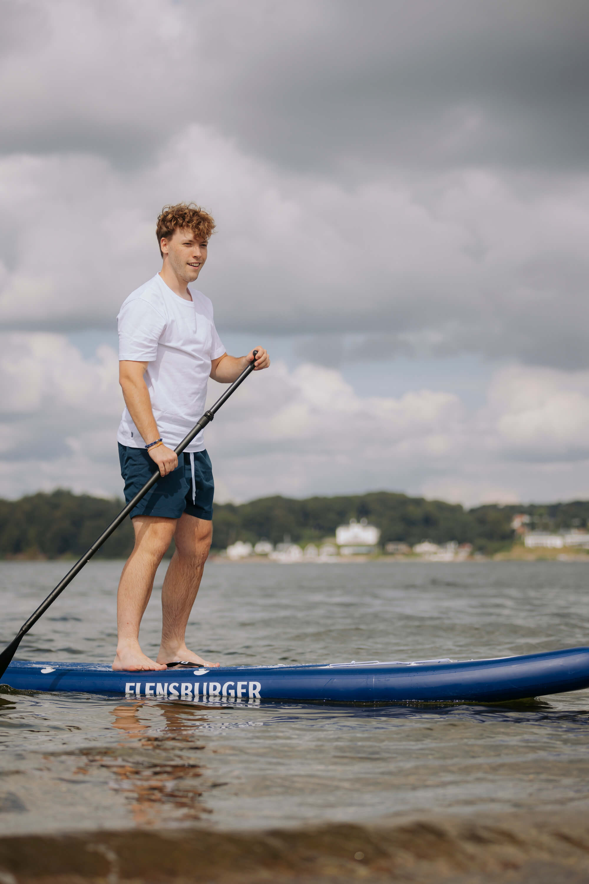 Ein junger Mann steht barfuß auf einem blauen SUP-Board mit seitlicher Aufschrift "FLENSBURGER" und hält ein Paddel. Er trägt ein weißes T-Shirt und blaue Shorts.