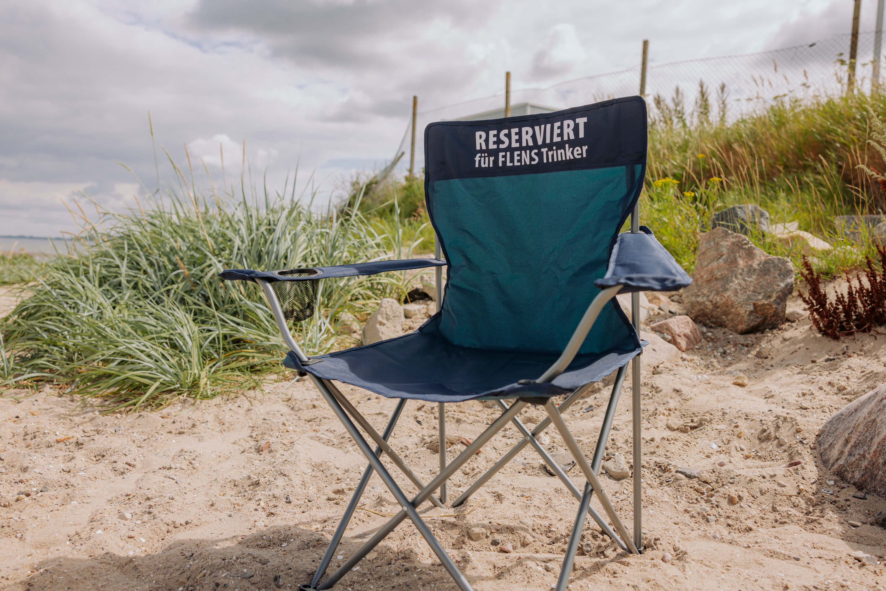 Campingstuhl "Fanø" steht am Strand. Auf der Rücklehne steht „RESERVIERT für FLENS Trinker“. Der Stuhl steht auf sandigem Boden, umgeben von hohem Gras und Steinen.