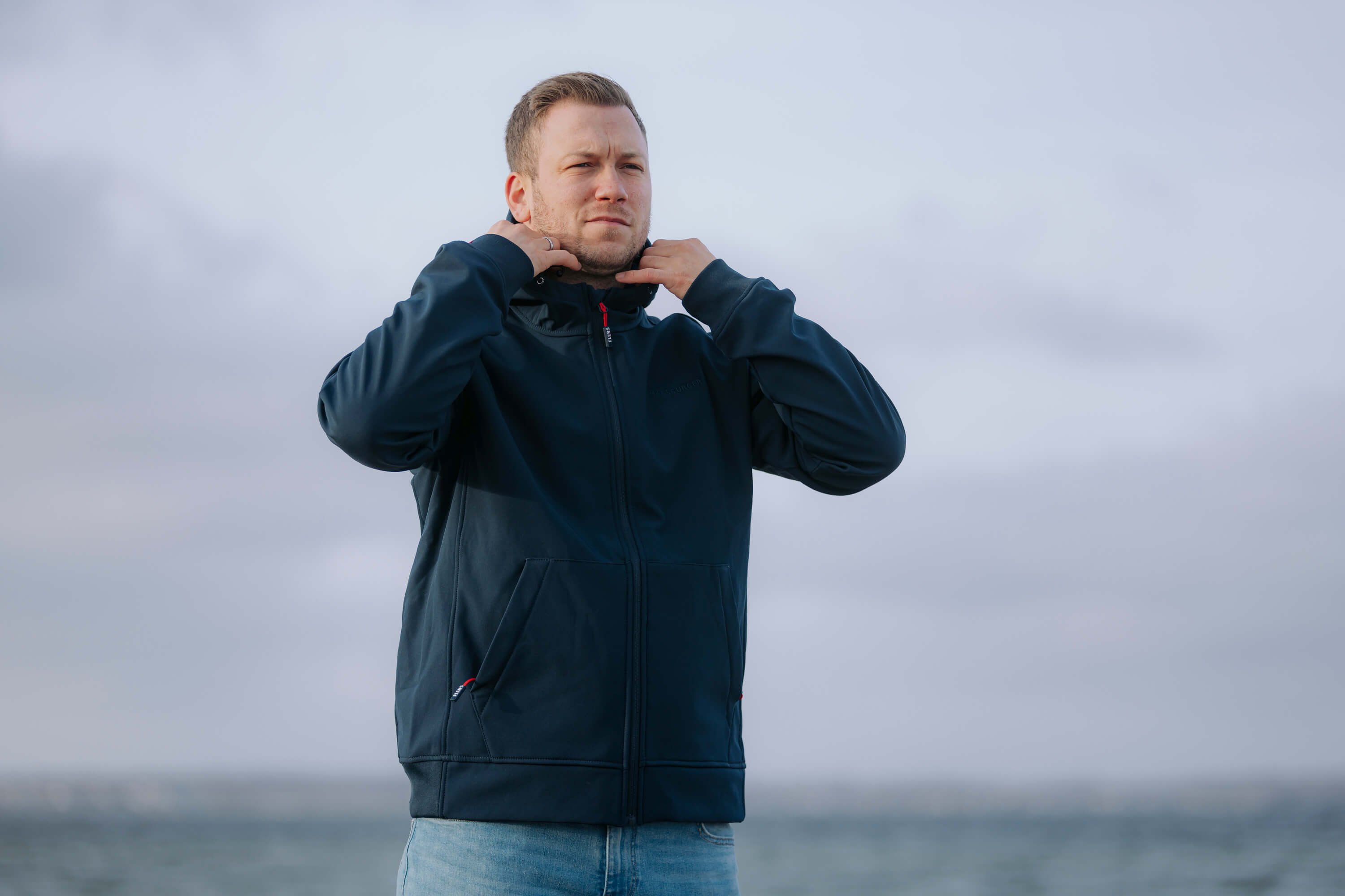 Mann trägt eine dunkelblaue Softshelljacke mit Reißverschluss und hält den Jackenkragen mit beiden Händen. Er steht vor einem bewölkten Himmel und einem Gewässer im Hintergrund.