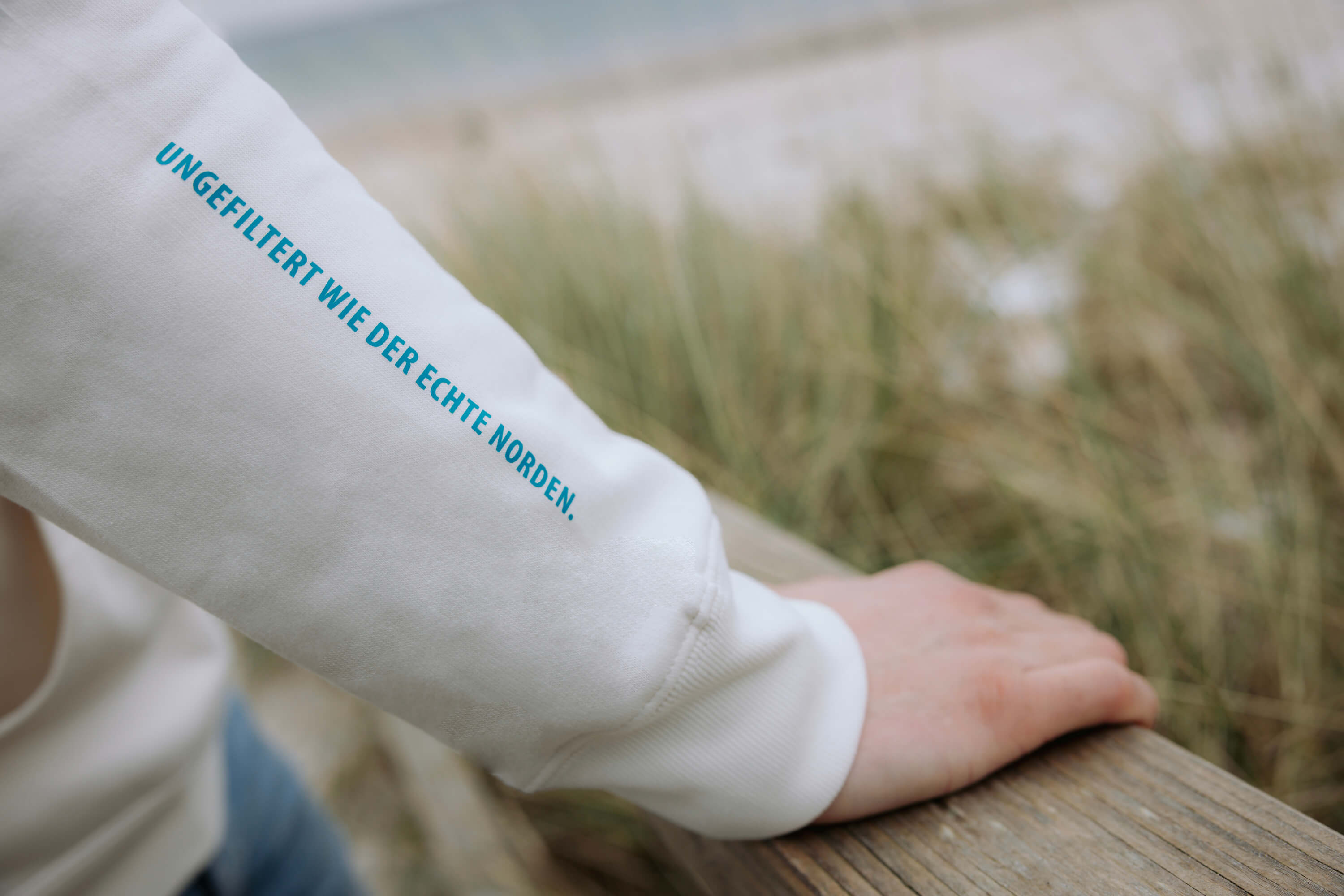 Detailansicht eines Hoodieärmels mit der blauen Aufschrift „Ungefiltert wie der echte Norden.“. Die aus dem Ärmel ragende Hand liegt auf einem Holzgeländer, im Hintergrund ist Gras und Strand zu sehen.