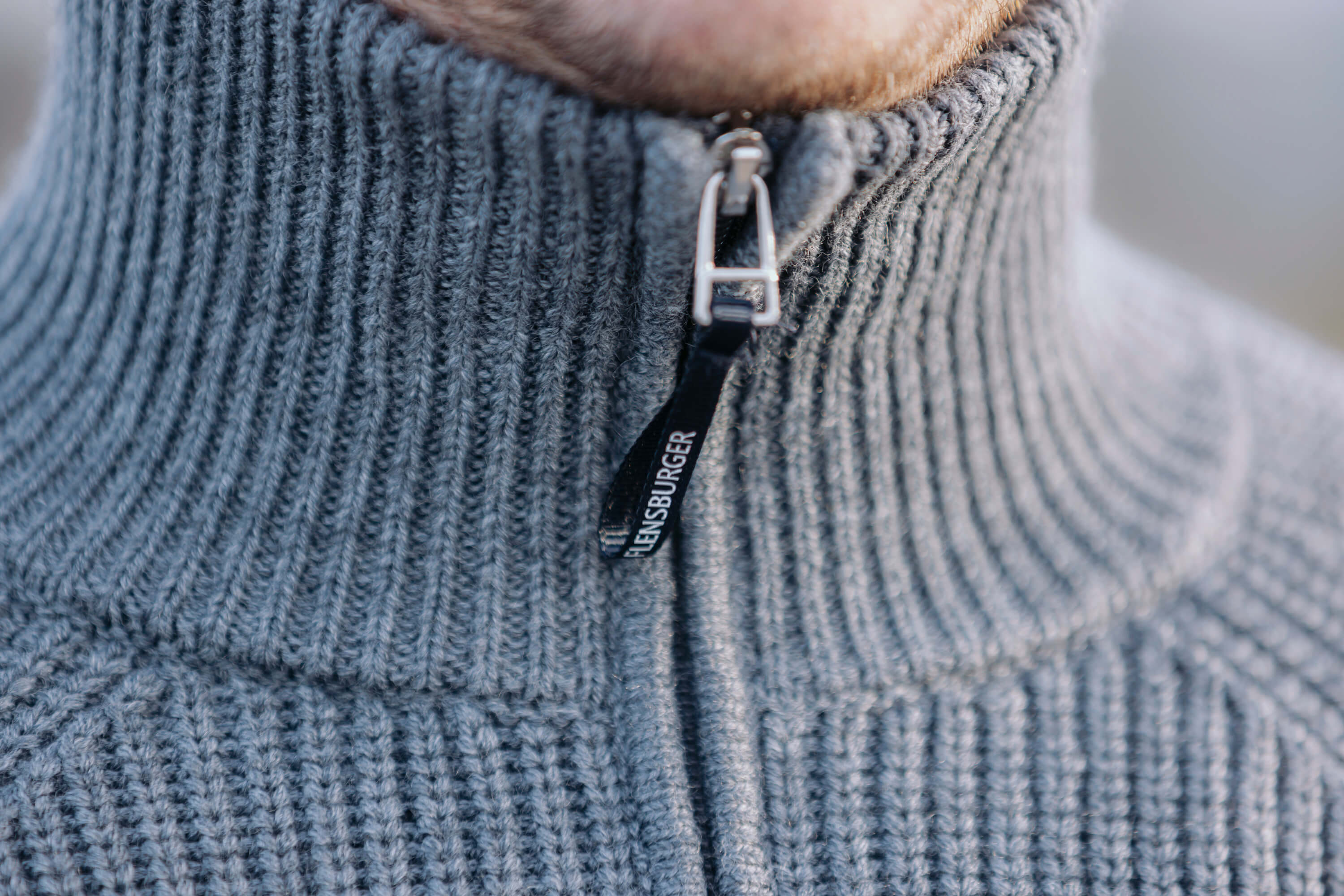 Detailansicht eines grauer, gerippter Strick-Troyer mit hohem Kragen und Reißverschluss. Am Reißverschluss hängt ein schwarzer Zipper-Anhänger mit weißer Aufschrift „HENSBURGER“.