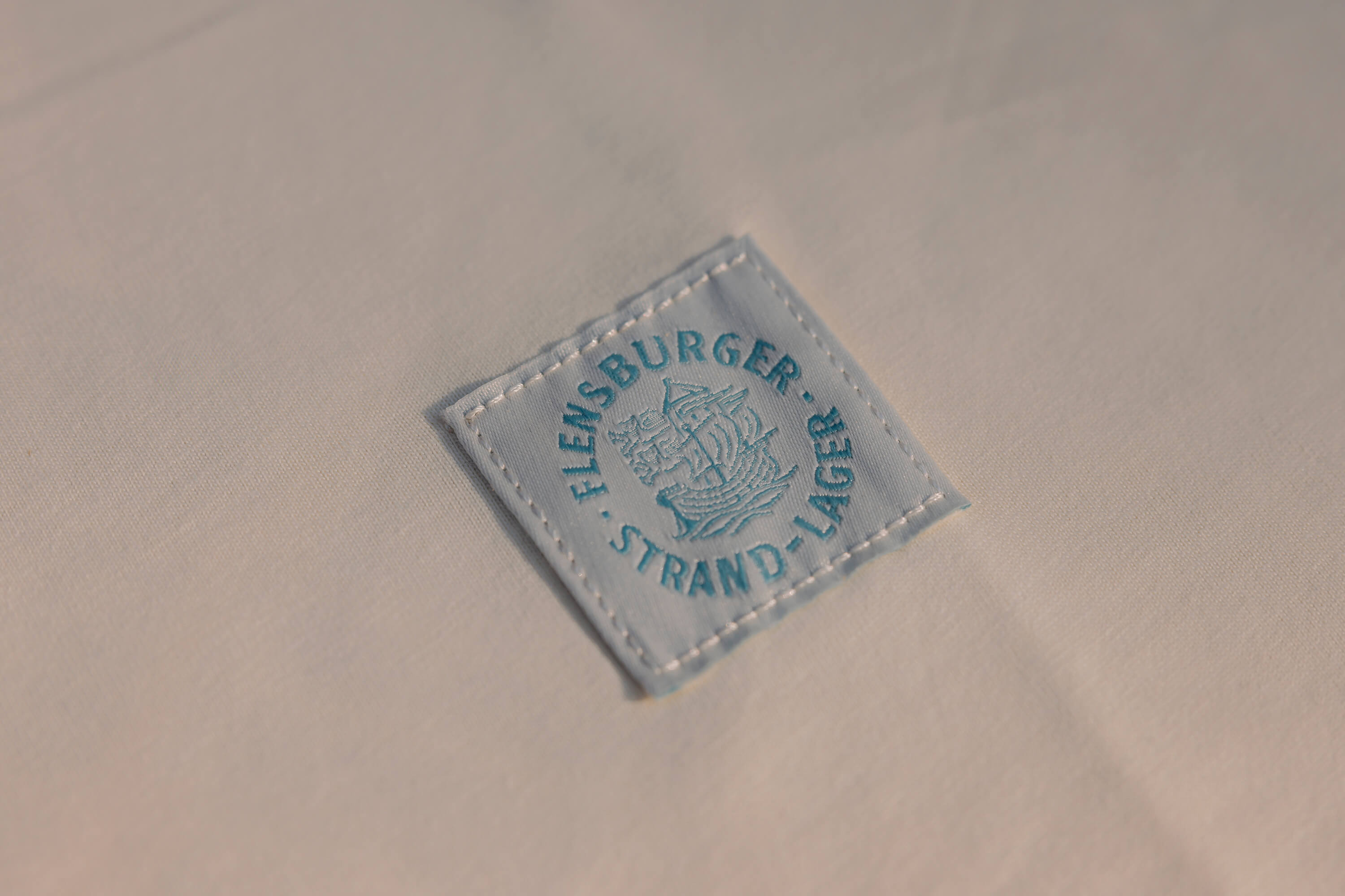 Weisses Textil mit einem weißen, eckigen Aufnäher. Auf dem Aufnäher ist ein türkises Segelschiff abgebildet und der Schriftzug „FLENSBURGER STRAND-LAGER“ steht rundherum.