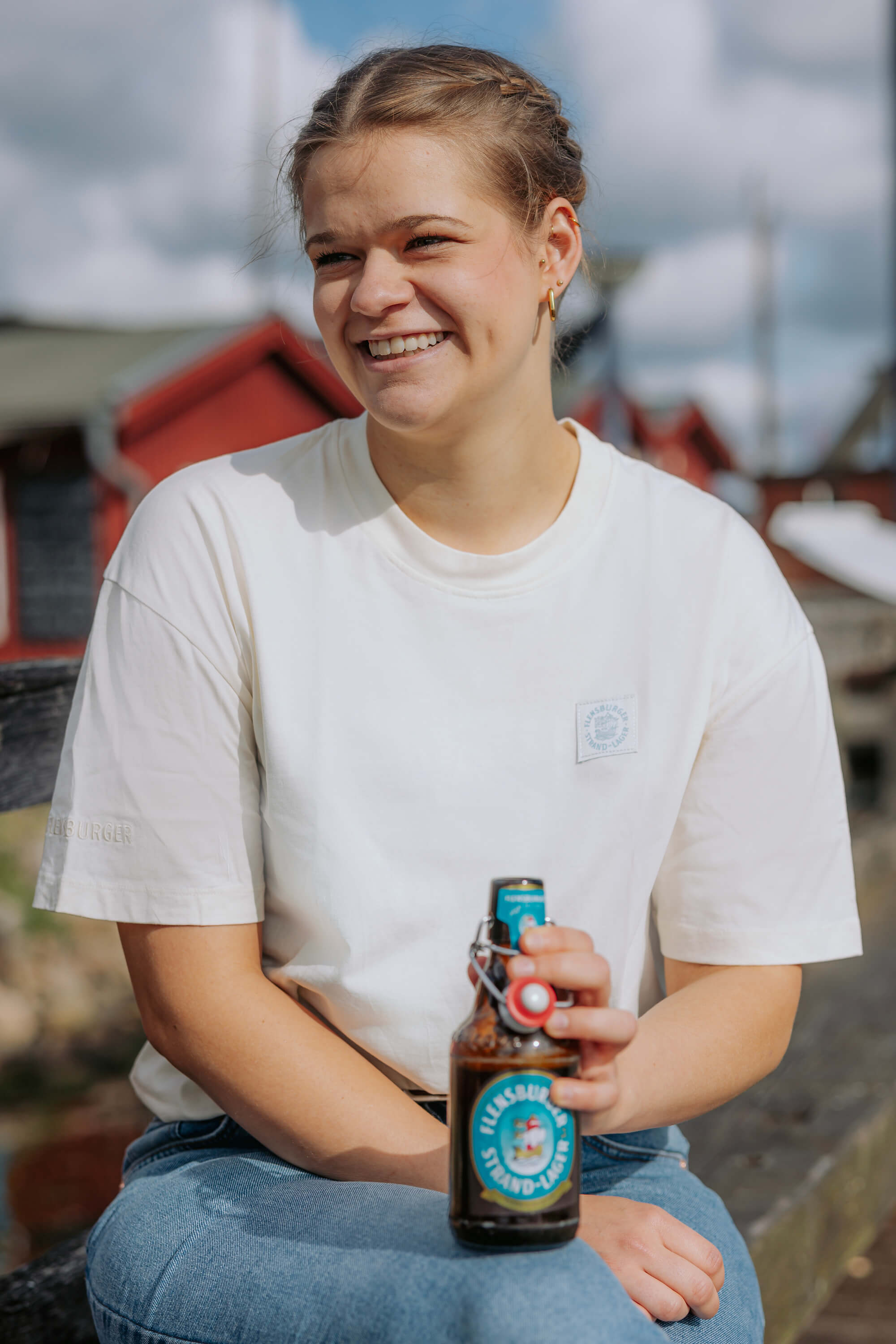 Junge Frau sitzt draußen auf einer Bank und hält eine Flasche Flensburger Strand-Lager in der Hand. Sie trägt ein weißes FLENS Strand-Lager T-Shirt aus 100% Bio-Baumwolle und lacht.