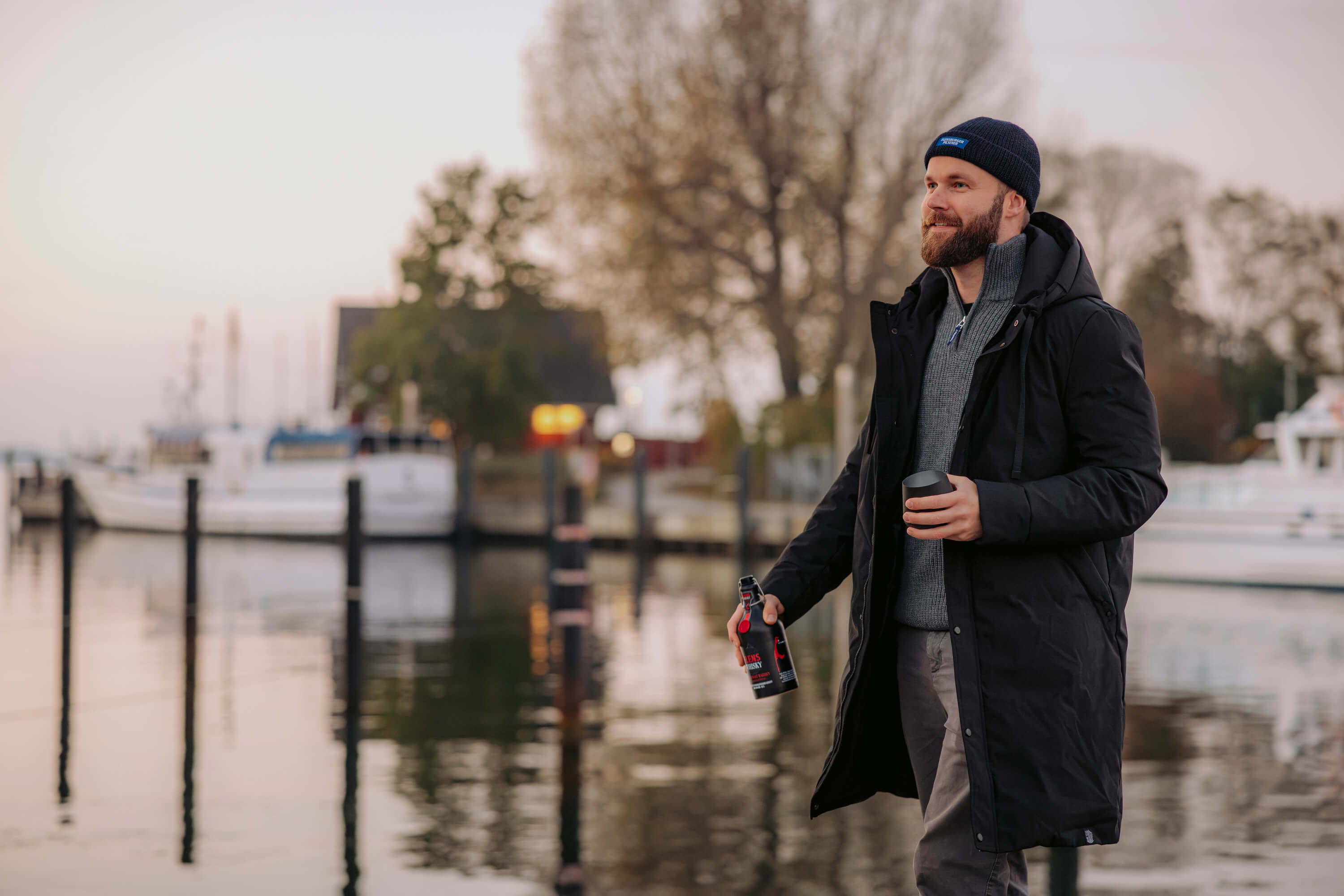 Mann trägt den schwarzen FLENS Winterparka "Ruben" mit Kapuze und Mütze offen an einem Hafen. Er hält eine schwarze Thermotasse in der linken und eine Flasche Flensburger Whisky in der rechten Hand.