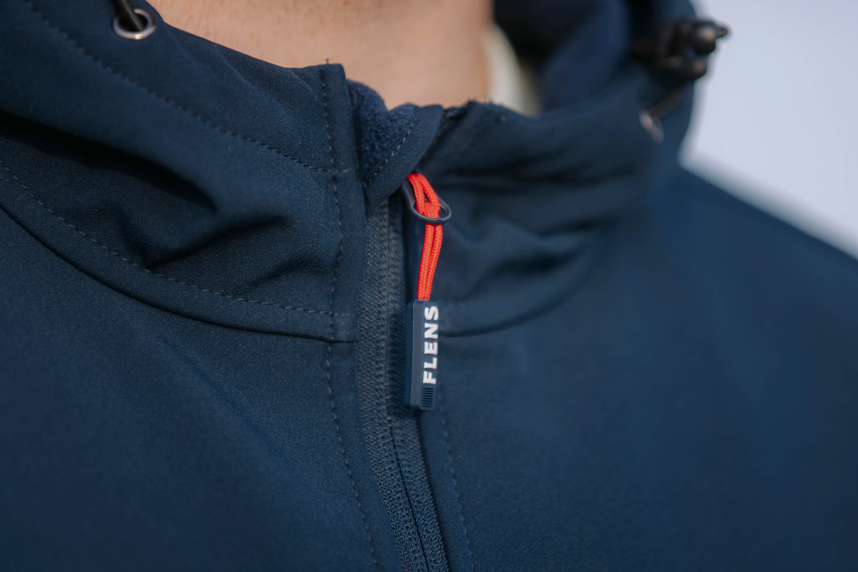 Detailansicht des Reißverschluss einer blauen Softshelljacke mit rotem Zipper-Anhänger, auf dem "FLENS" steht.