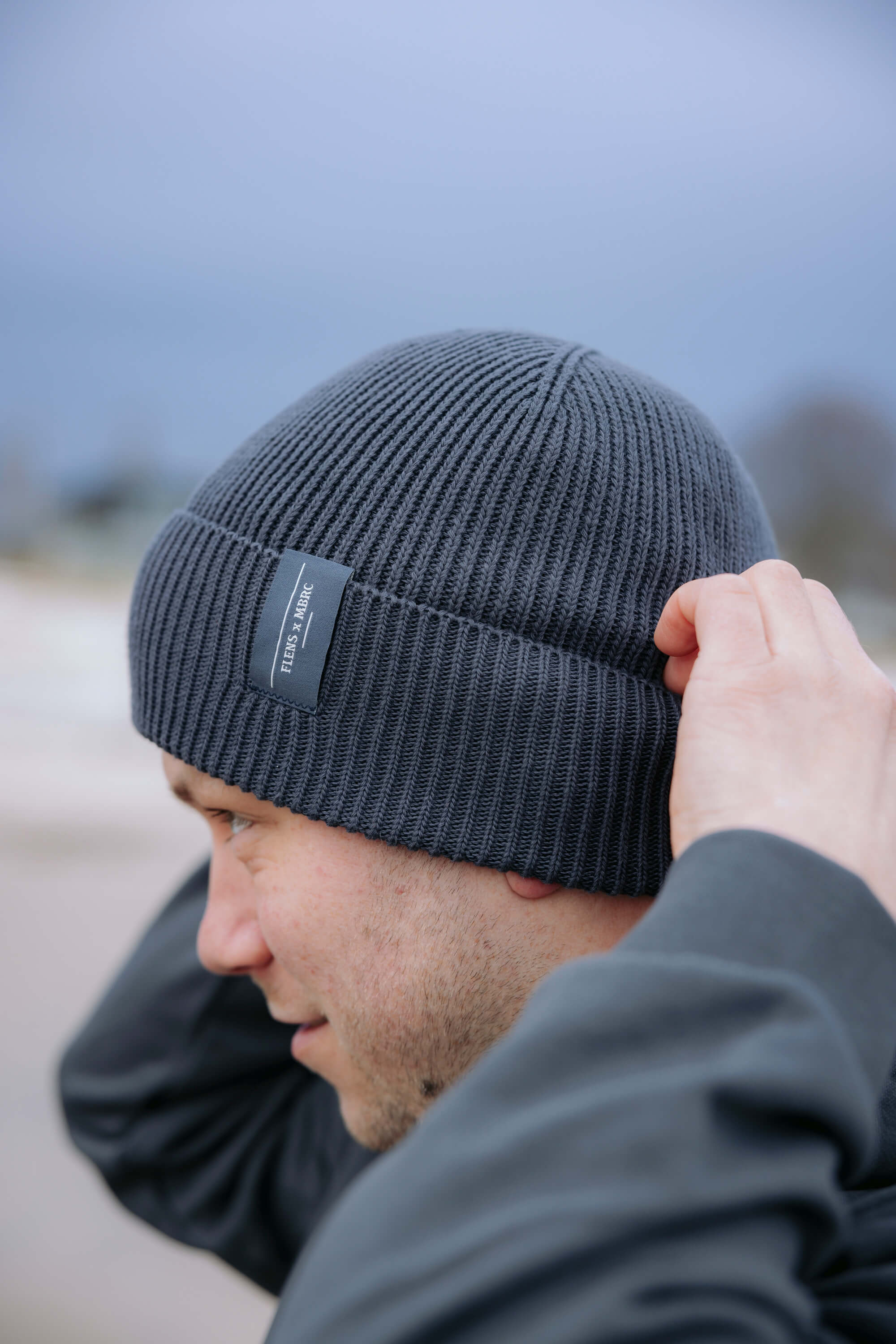 Schwarze, gerippte Bio-Baumwoll-Beanie mit umgeschlagenem Saum trägt das Logo „FLENS x MBRC“ in grauer Schrift. Eine Person zieht die Beanie locker über den Kopf.
