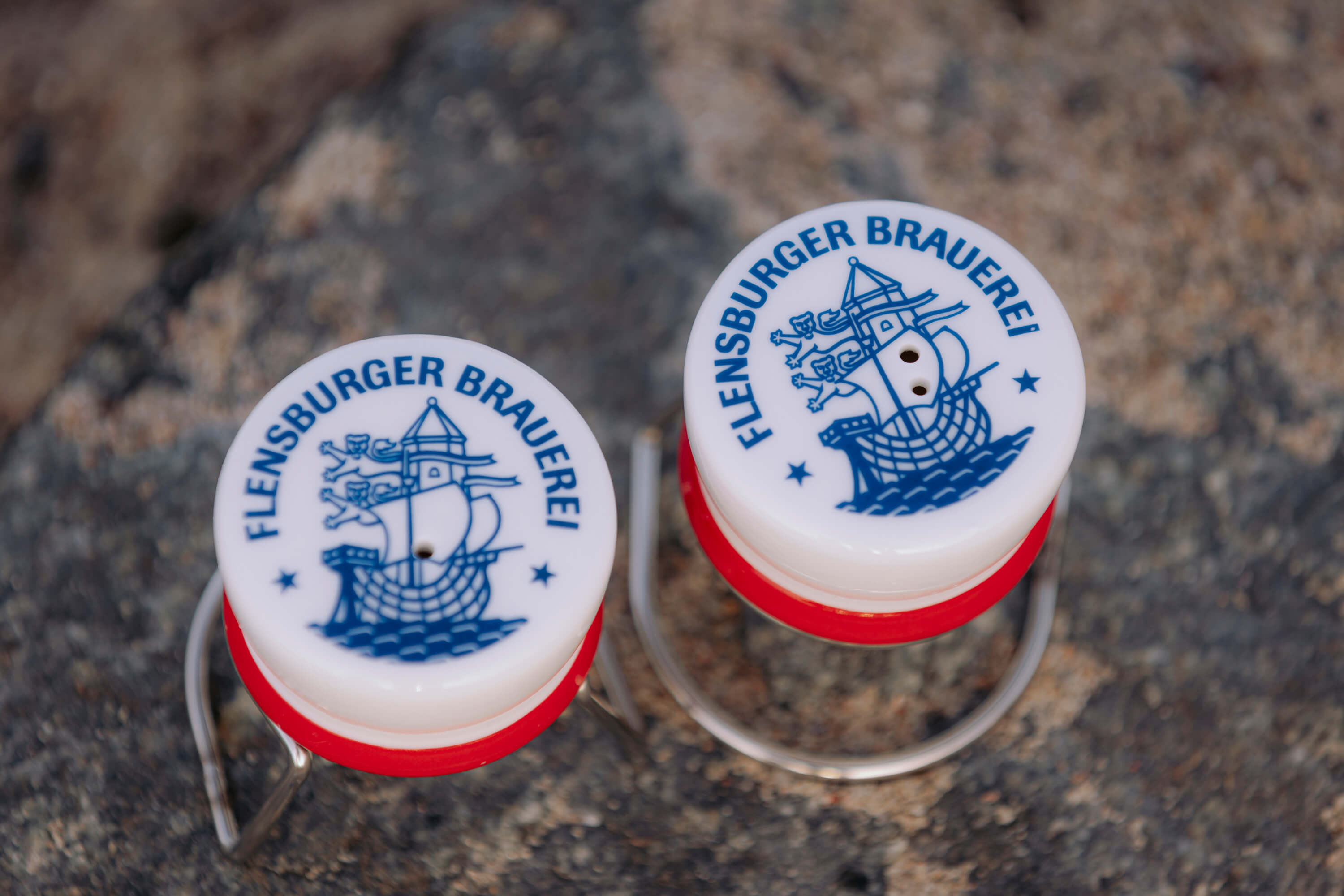 Salt- und Pfefferstreuer in Bügelverschlussoptik aus der Vogelperspektive. Zu sehen ist in dunkelblau die Aufschrift „Flensburger Brauerei“ und einem Schiff-Logo. Die beiden Streuer stehen auf einem felsigen Untergrund.