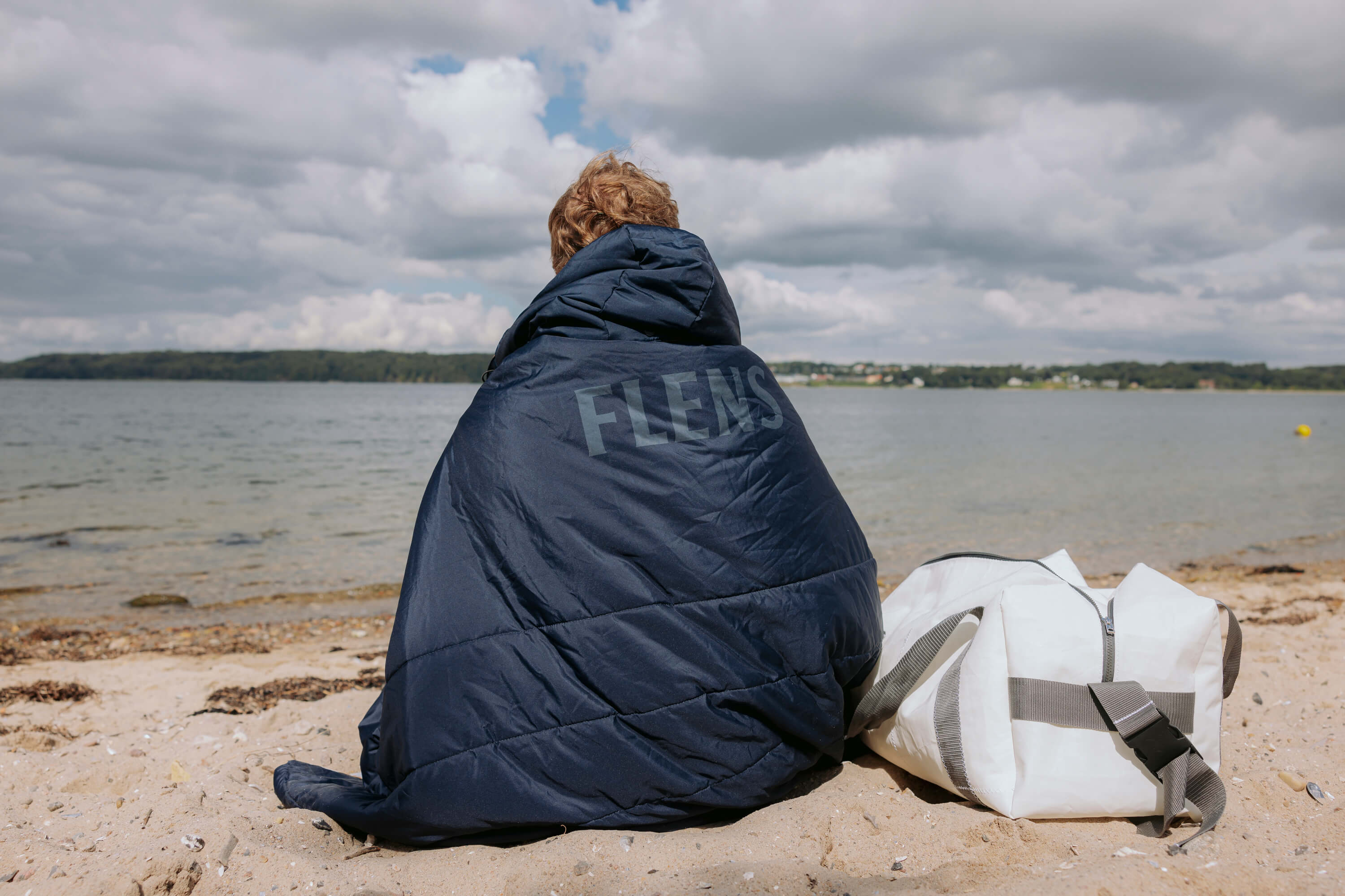 Person sitzt am Strand und hat sich eine dunkelblaue 3in1 Outdoordecke mit dem Aufdruck „FLENS“ über die Schultern gelegt. Daneben liegt eine weiße Tasche im Sand.