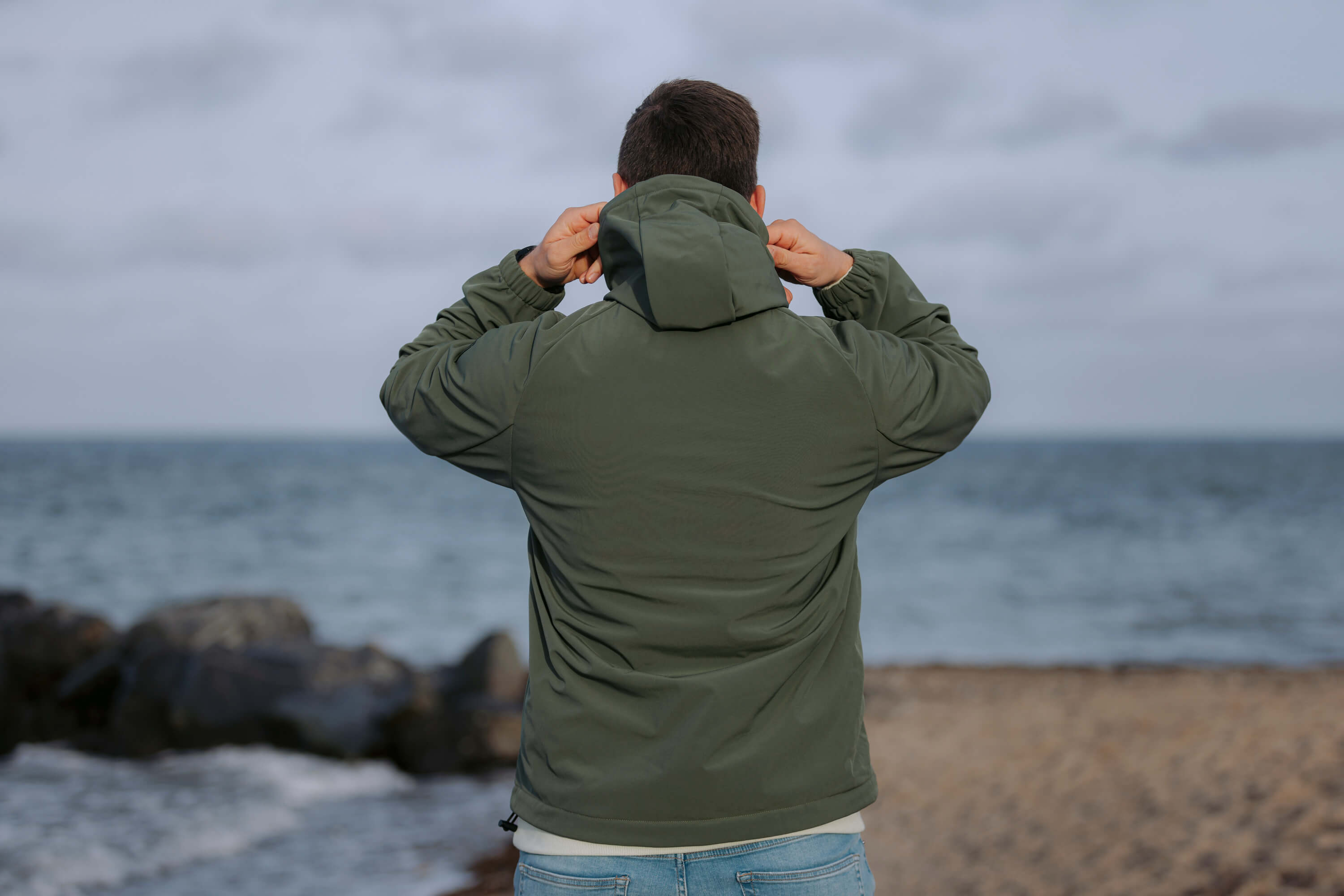 Ein Mann steht mit dem Rücken zur Kamera und zieht die Kapuze seines olivgrünen Windbreakers Skagerrak über den Kopf. Im Hintergrund sind Meer und Sandstrand zu sehen.