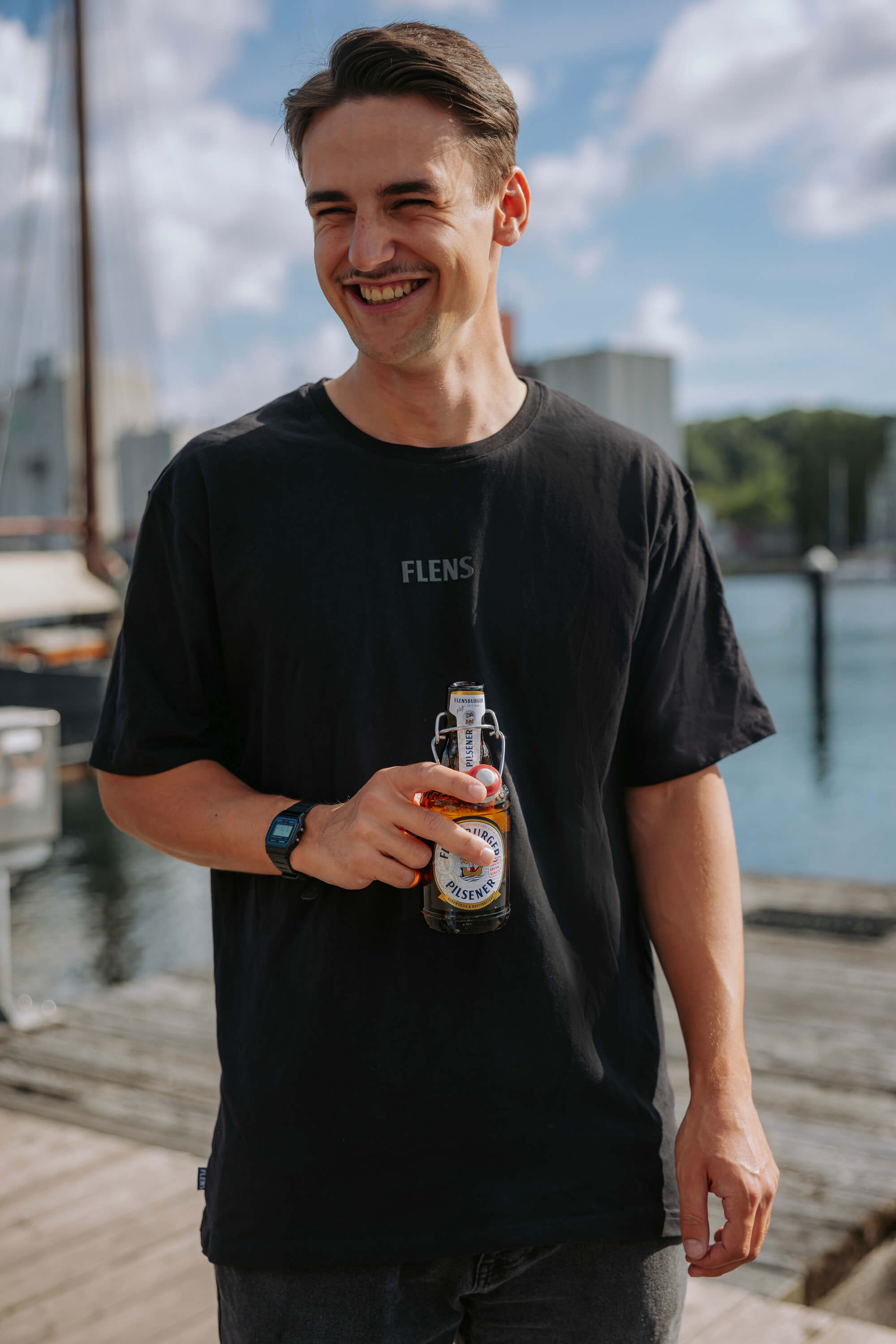 Junger Mann trägt ein anthrazitfarbenes T-Shirt mit der Aufschrift "FLENS" mittig auf der Brust. Er hält eine Flasche Flensburger Pilsener und steht an einem Hafen.